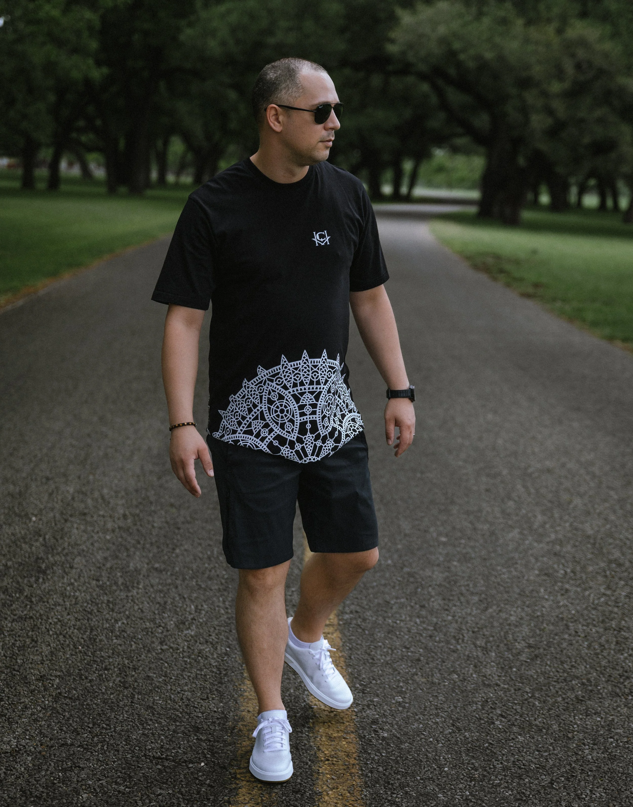 A man wearing sunglasses, a black t-shirt with a blue emblem, black shorts, and white sneakers, walking on an asphalt road in a park with green trees in the background.