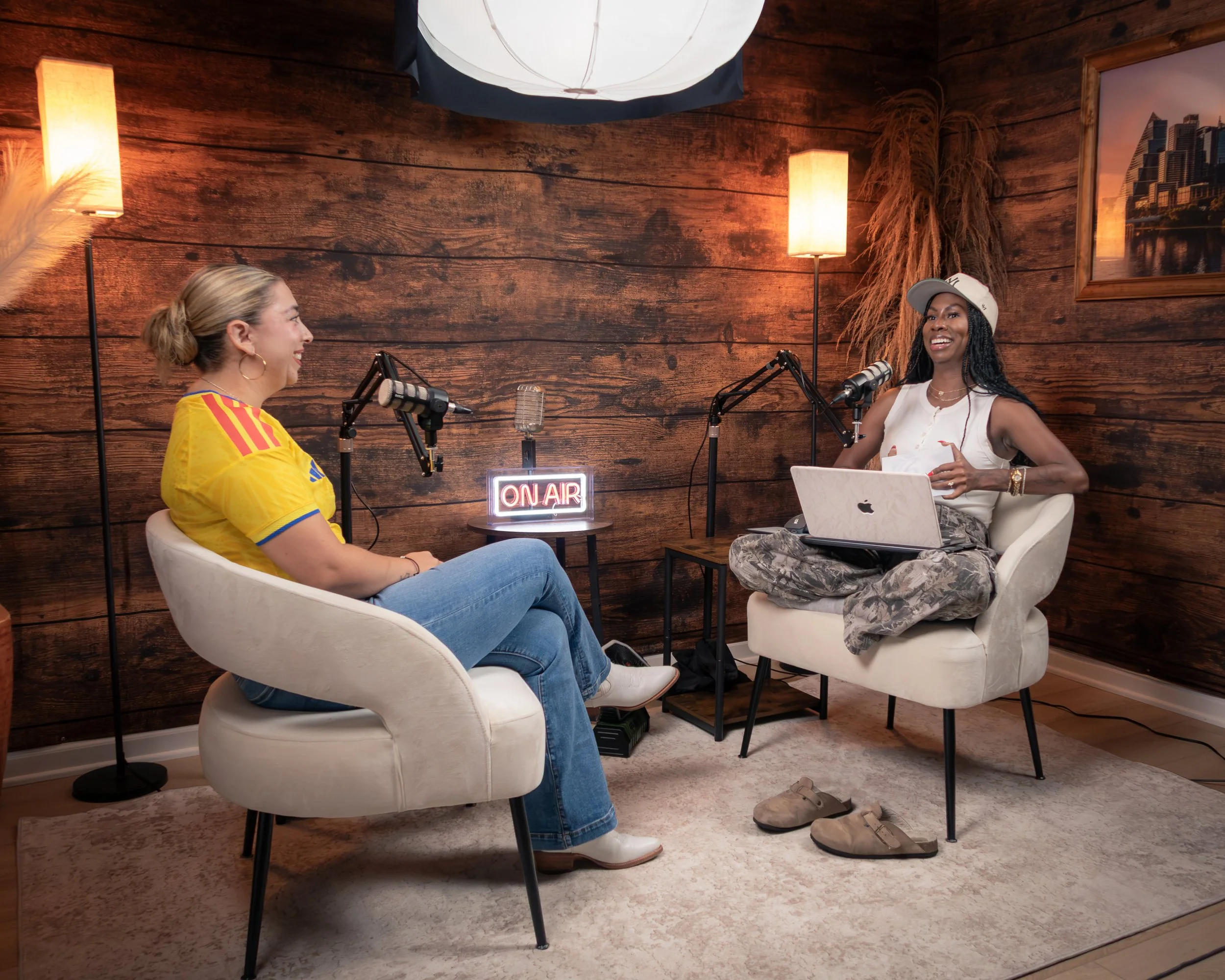 Two women having a podcast conversation in a cozy room with wooden walls, recorded with microphones. One woman is wearing a yellow sports jersey and the other a white sleeveless top and a white cap. There is an 'On Air' sign between them, a framed cityscape photo on the wall, and warm lighting from two floor lamps.