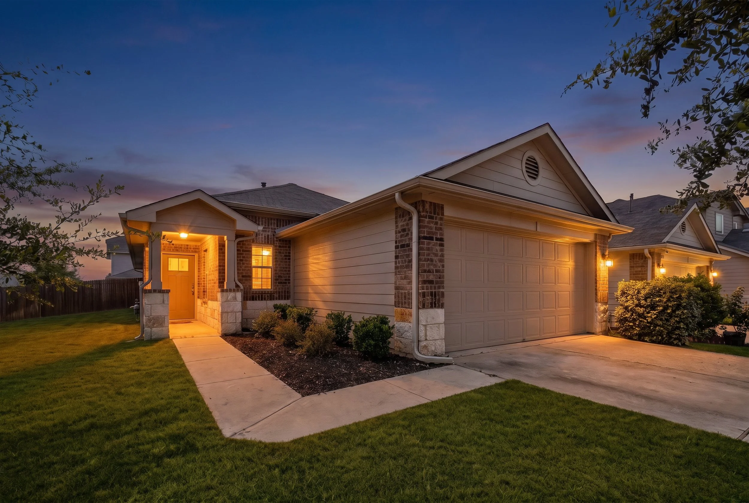 Front view of a suburban house at dusk with warm exterior lighting, a two-car garage, a brick and siding exterior, a manicured lawn, and a sidewalk leading to the front door.