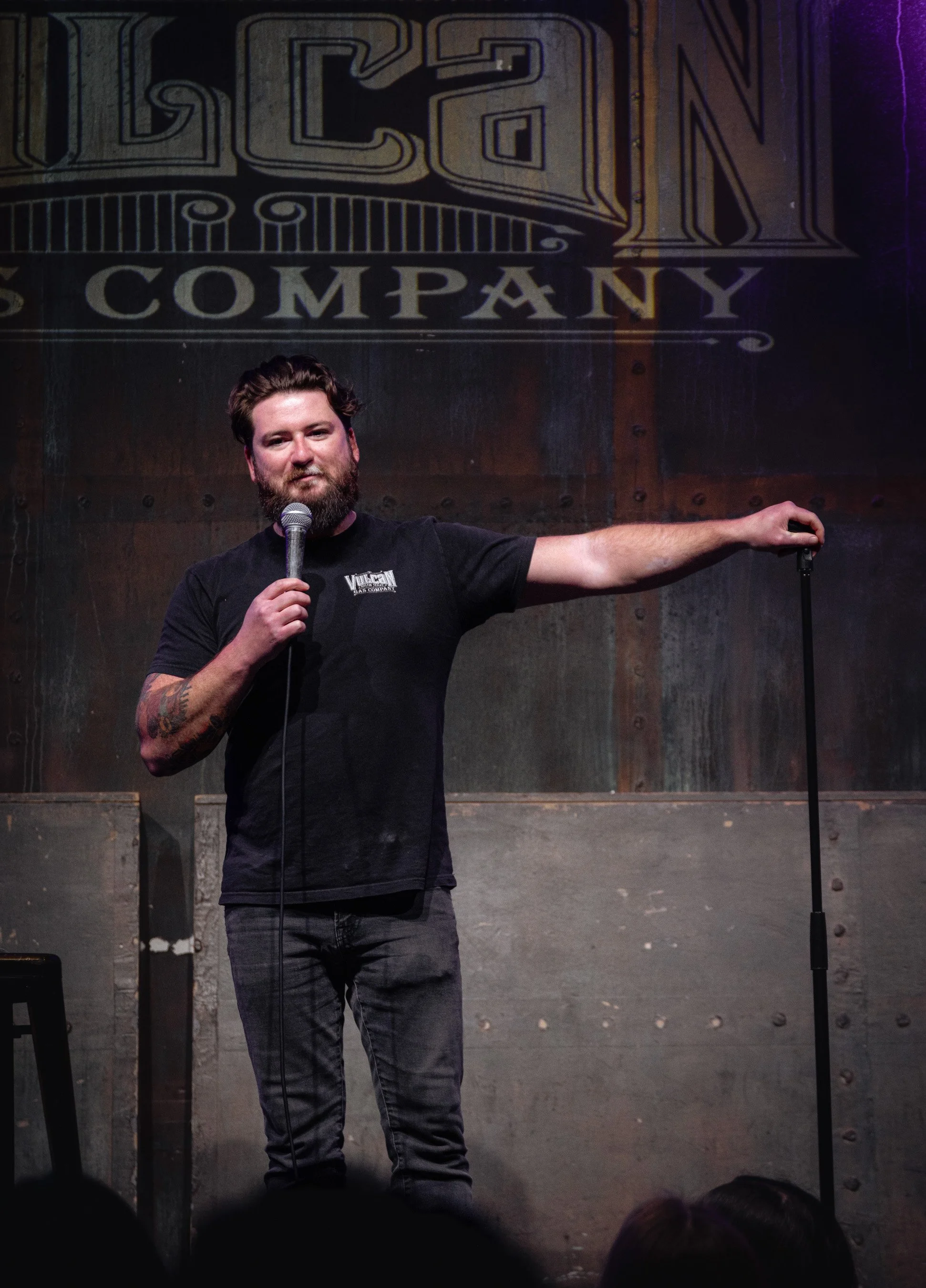 A man with a beard and tattoos on his arm holding a microphone and pointing to the right on a stage with a metal and wood backdrop featuring a sign that reads 'STONE COMPANY'.