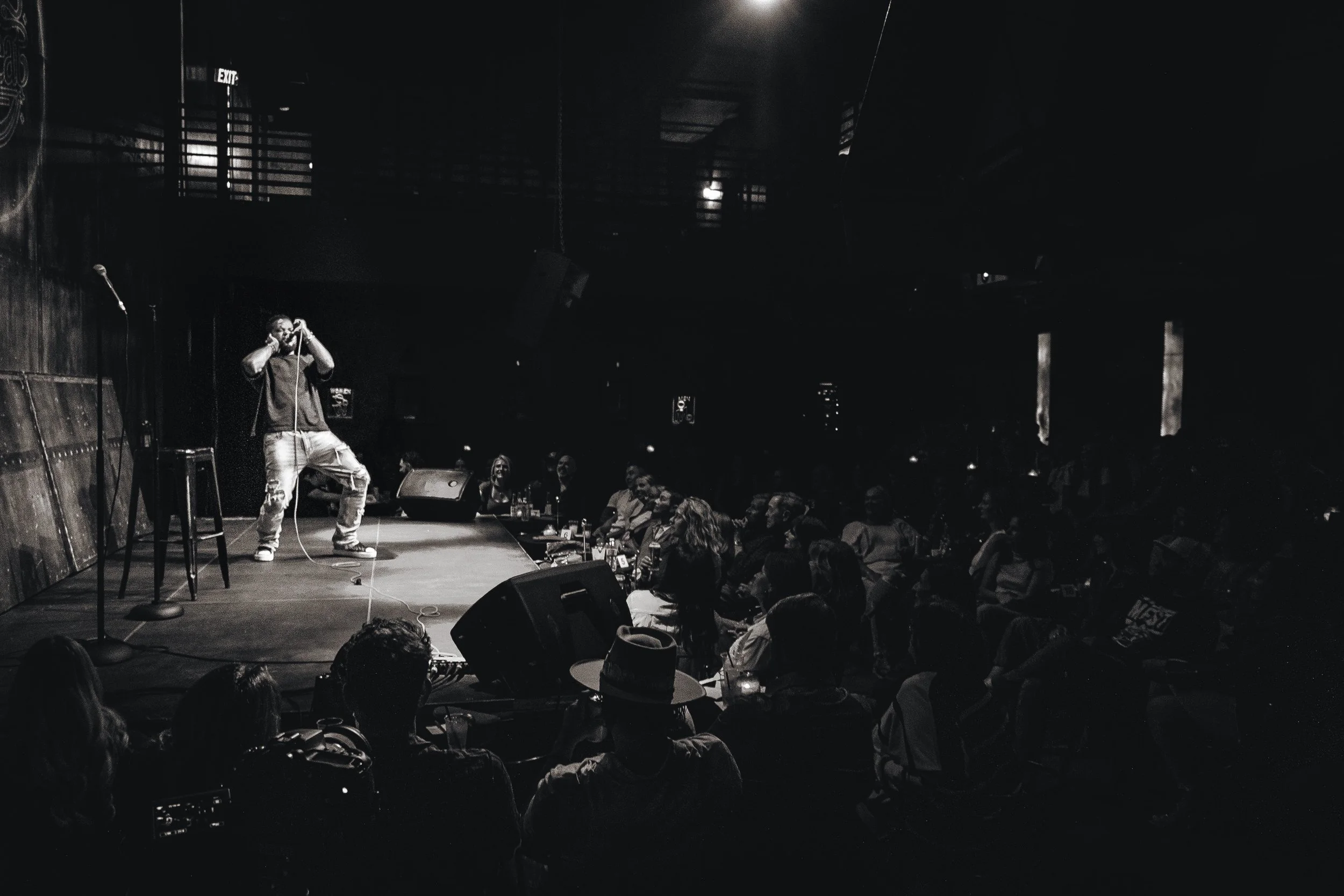 A comedian performs on stage at a comedy club, with an audience seated at tables in the foreground. The scene is dark with focused lighting on the performer, who is holding a microphone and standing in front of a black background.