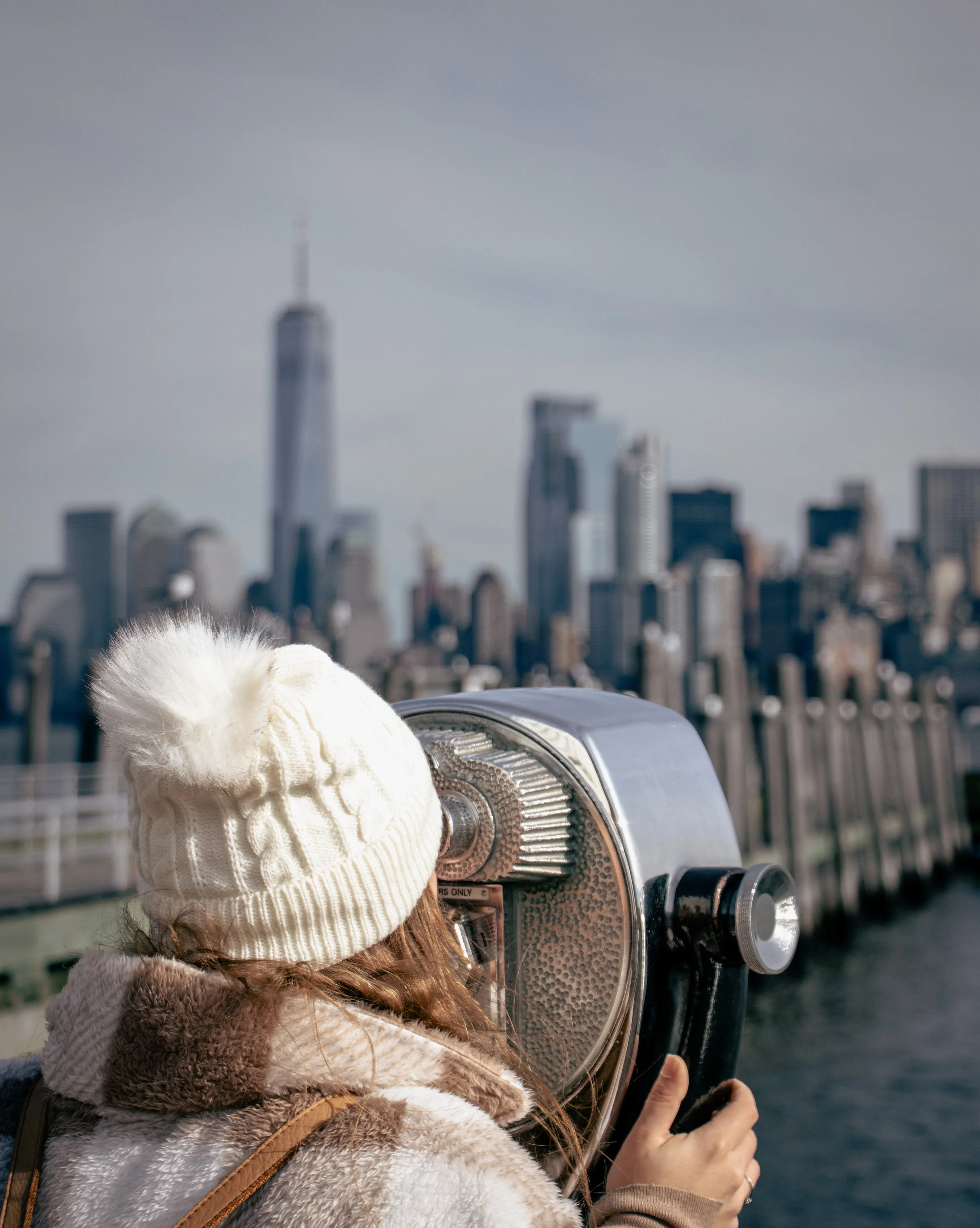 A woman with long reddish-brown hair wearing a cream-colored knit hat and a tan faux fur coat, looking through a coin-operated binocular viewer on a rooftop with a city skyline in the background.