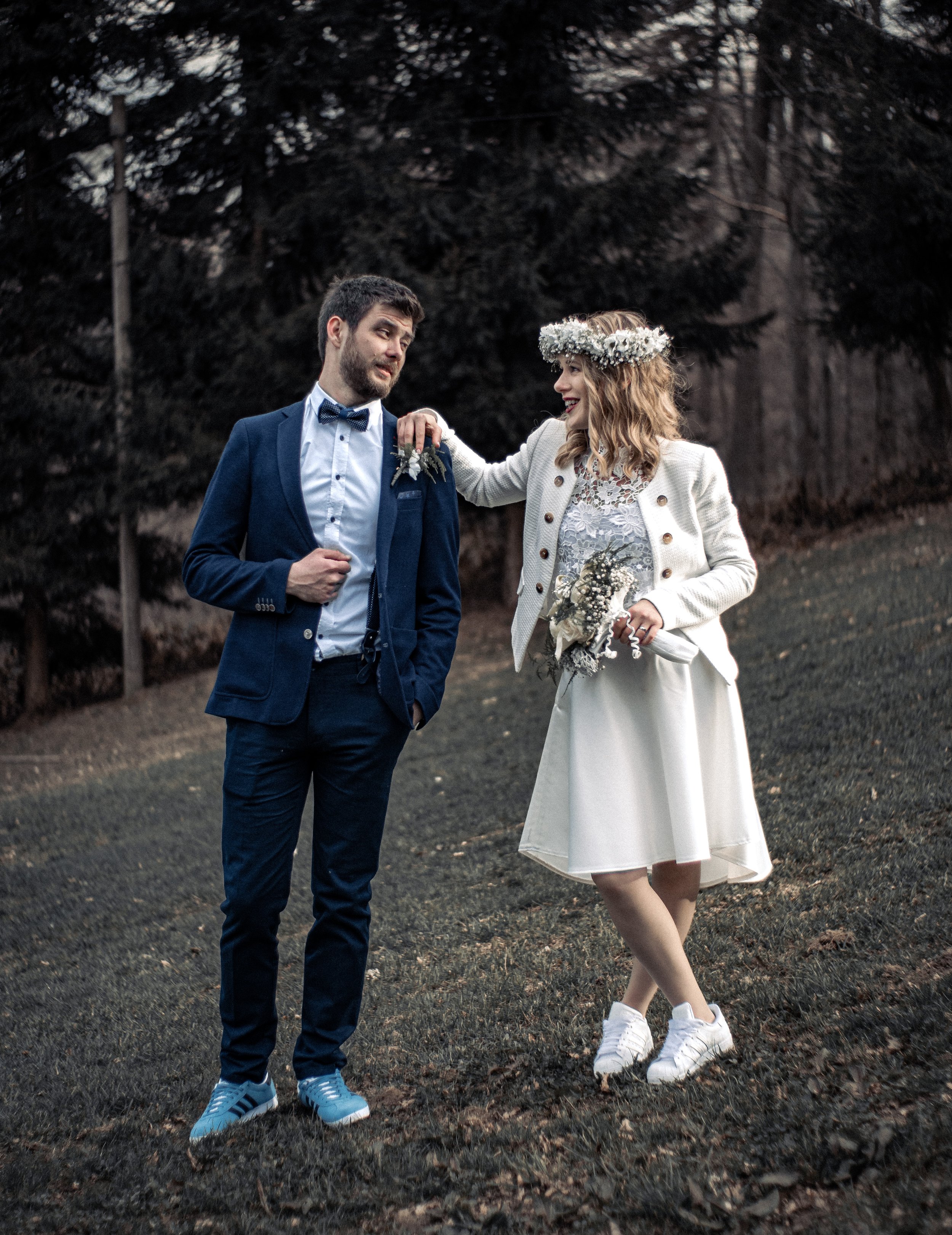 A man in a blue suit and a woman in a white dress with a floral crown are standing outdoors on a grassy area with trees in the background. The woman is smiling, holding a bouquet, and touching the man's shoulder.