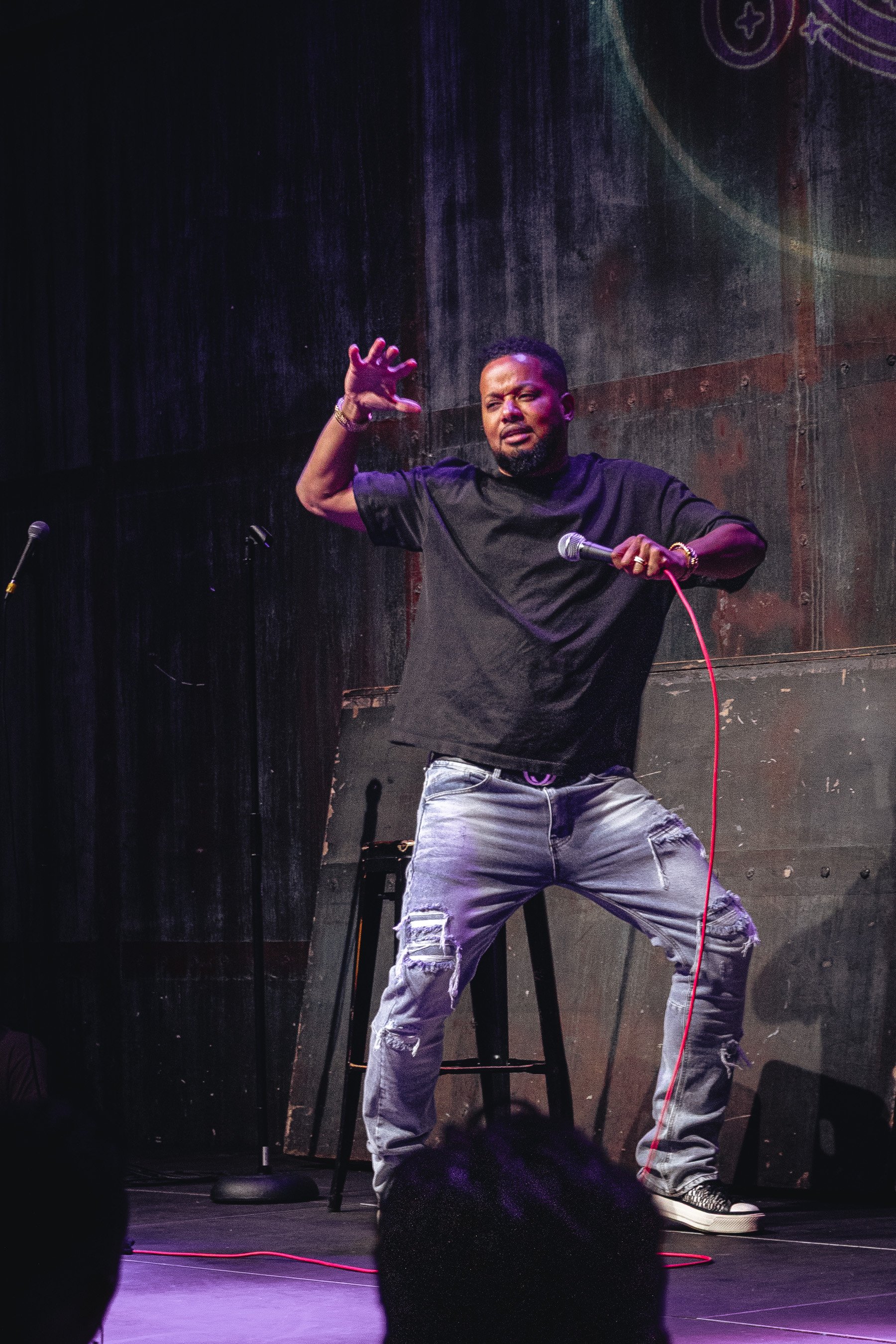 A man performing stand-up comedy on stage, holding a microphone in his right hand while gesturing with his left hand. He is wearing a black t-shirt, distressed jeans, and sneakers. The background features a dark wall with a textured surface.