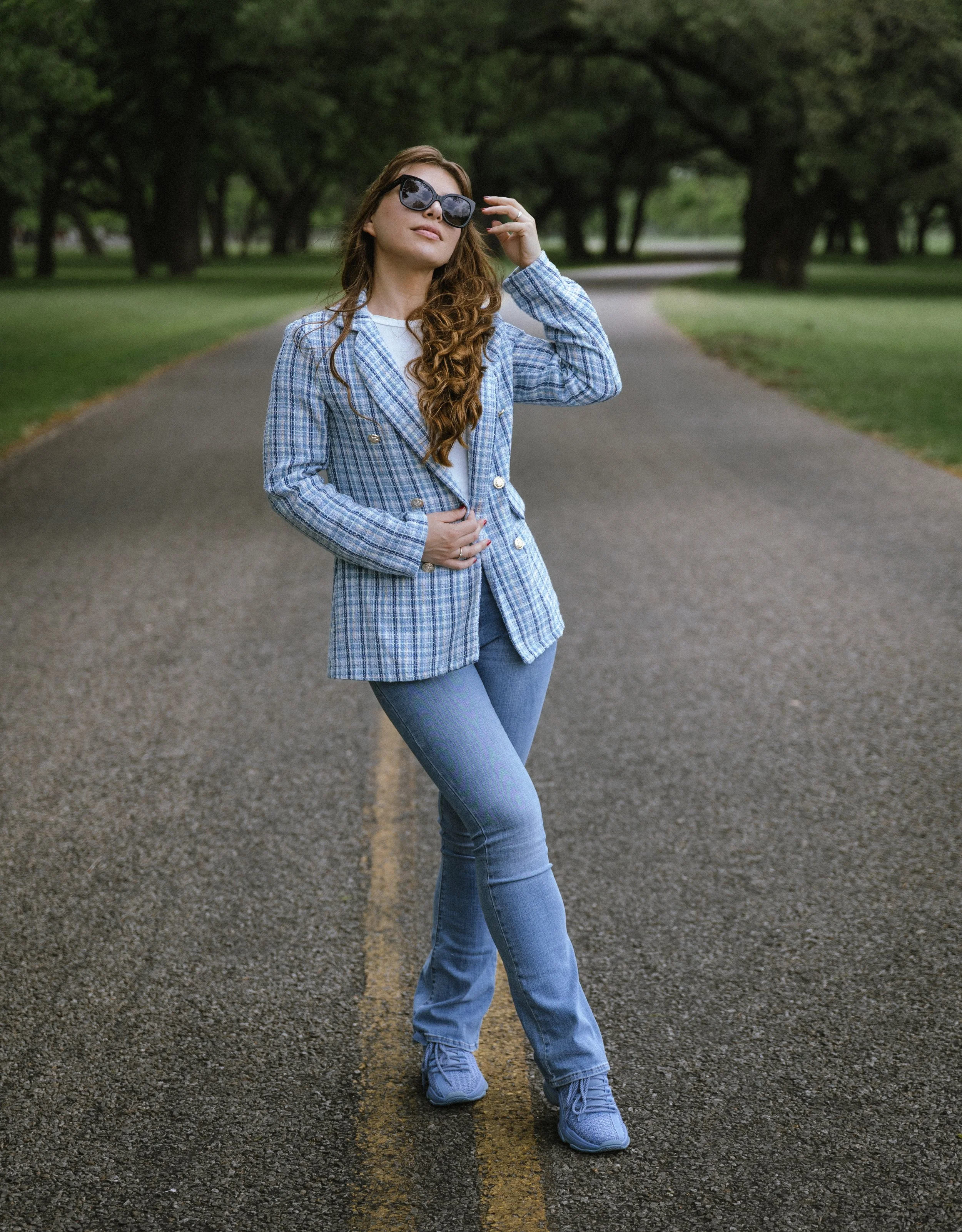 Young woman standing on a tree-lined rural road, wearing sunglasses, a blue plaid blazer, jeans, and sneakers, with a confident pose.
