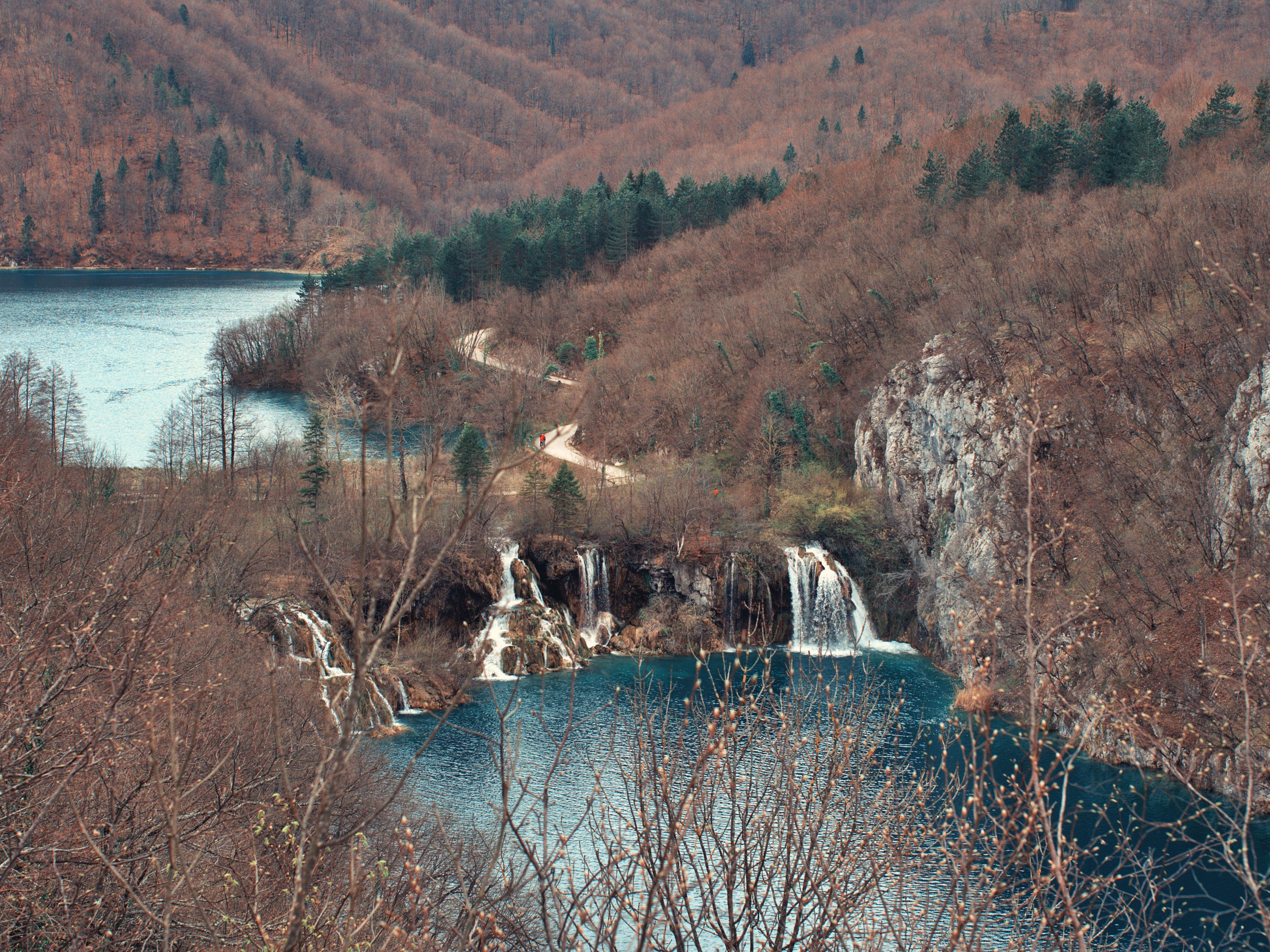 A scenic view of a mountainous landscape with a waterfall cascading into a blue lake, surrounded by leafless trees and forested hills.