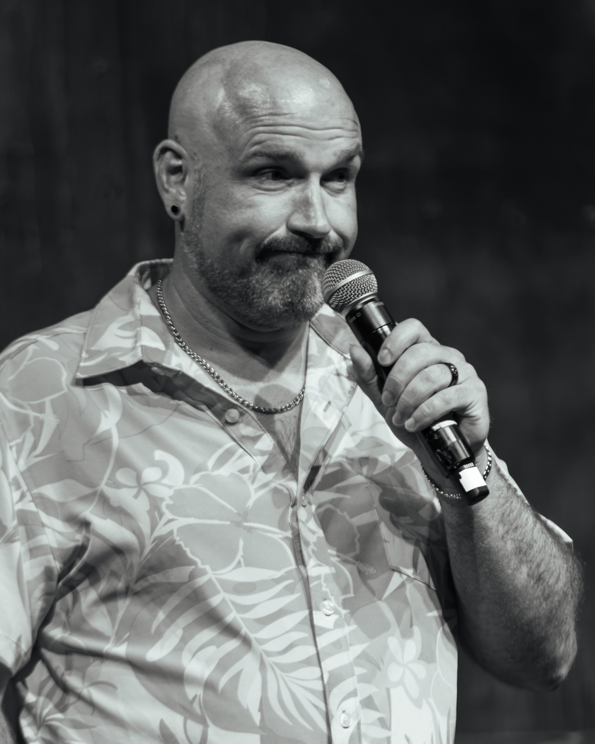 A man speaking into a microphone, with a shaved head, beard, and wearing earrings, a patterned shirt, and a chain necklace, against a dark background.