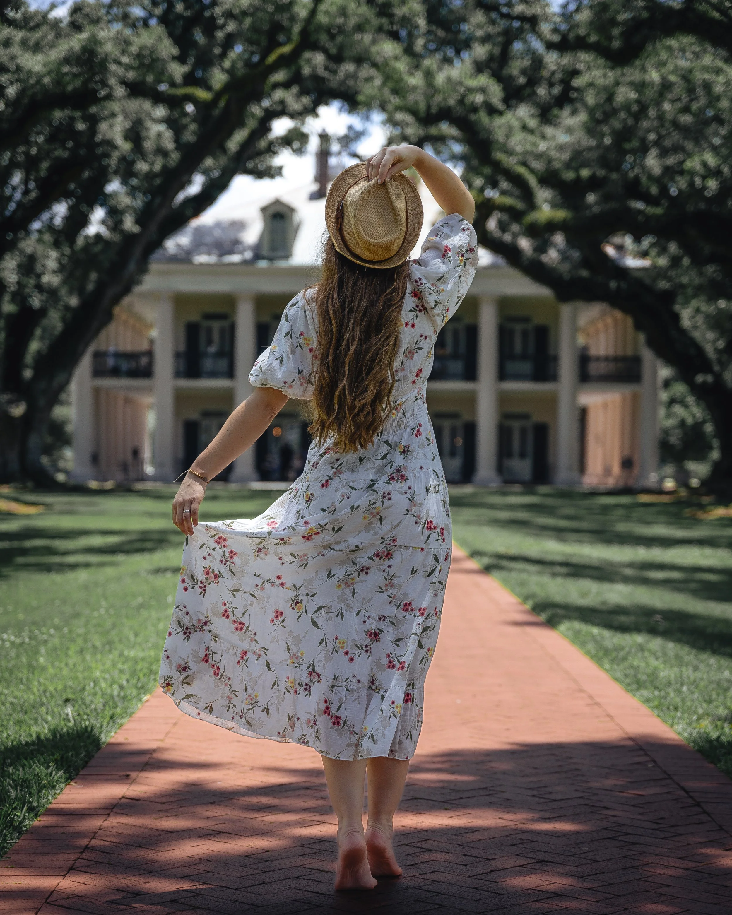 A woman with long hair wearing a floral dress, holding a straw hat, standing barefoot on a brick pathway under large trees with a building in the background.