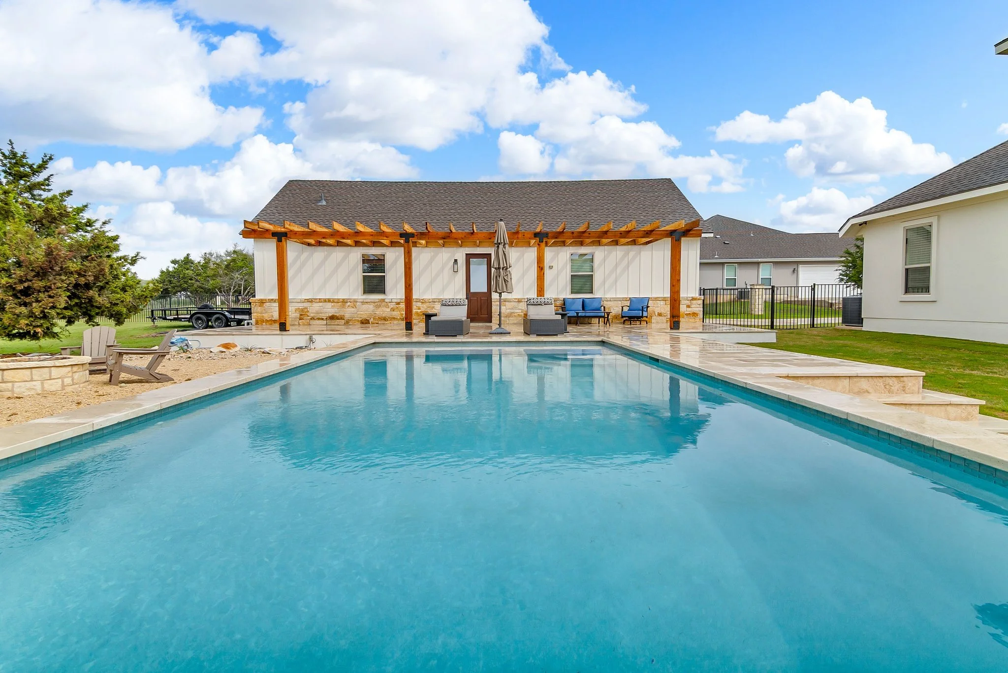 Residential backyard with a swimming pool in the foreground, patio furniture including chairs and an umbrella on a deck, a house with an open pergola, and neighboring houses under a partly cloudy sky.