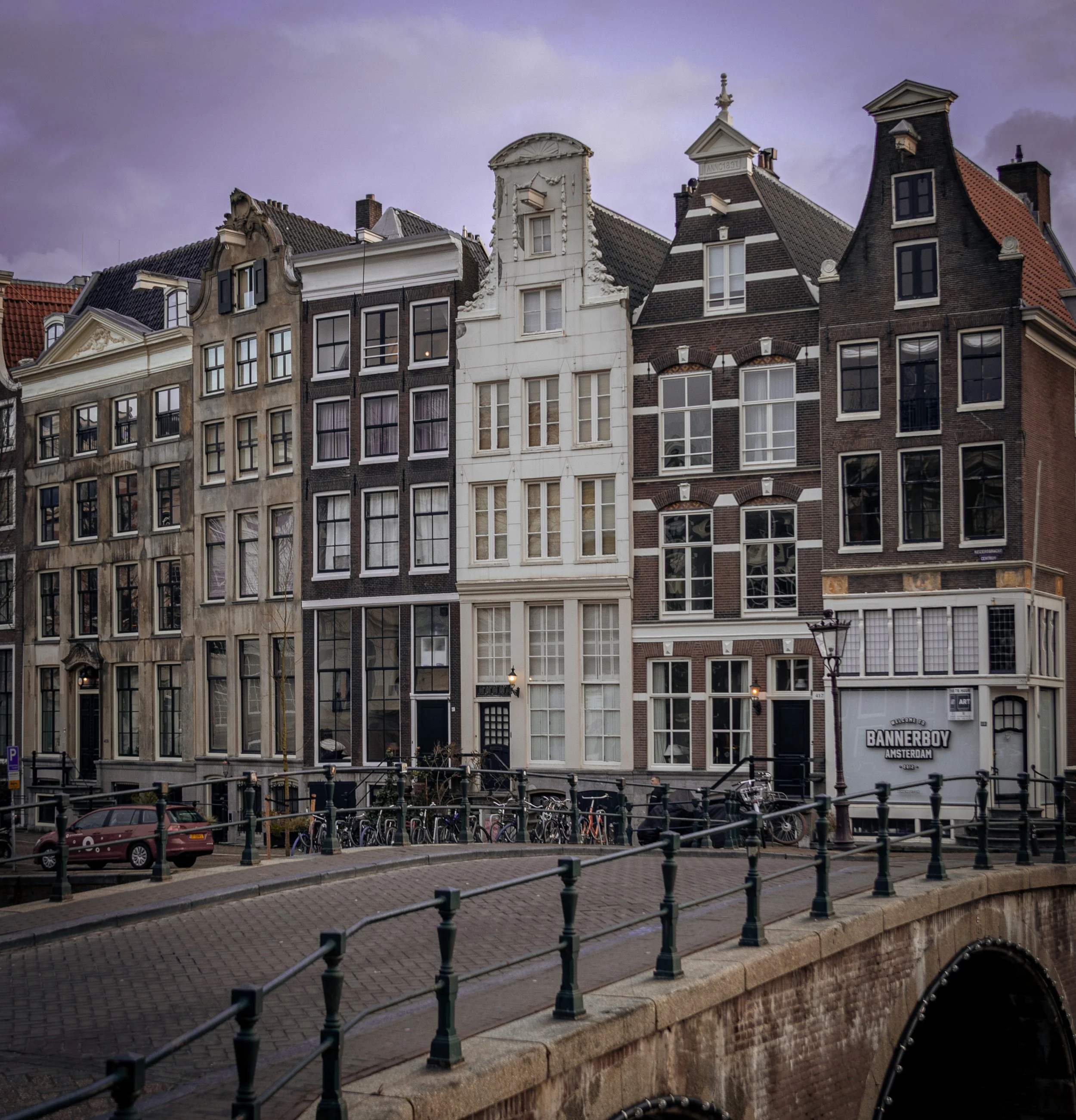 View of several narrow, historic buildings with classic Dutch gabled facades along a canal in Amsterdam, with a purple cloudy sky overhead.