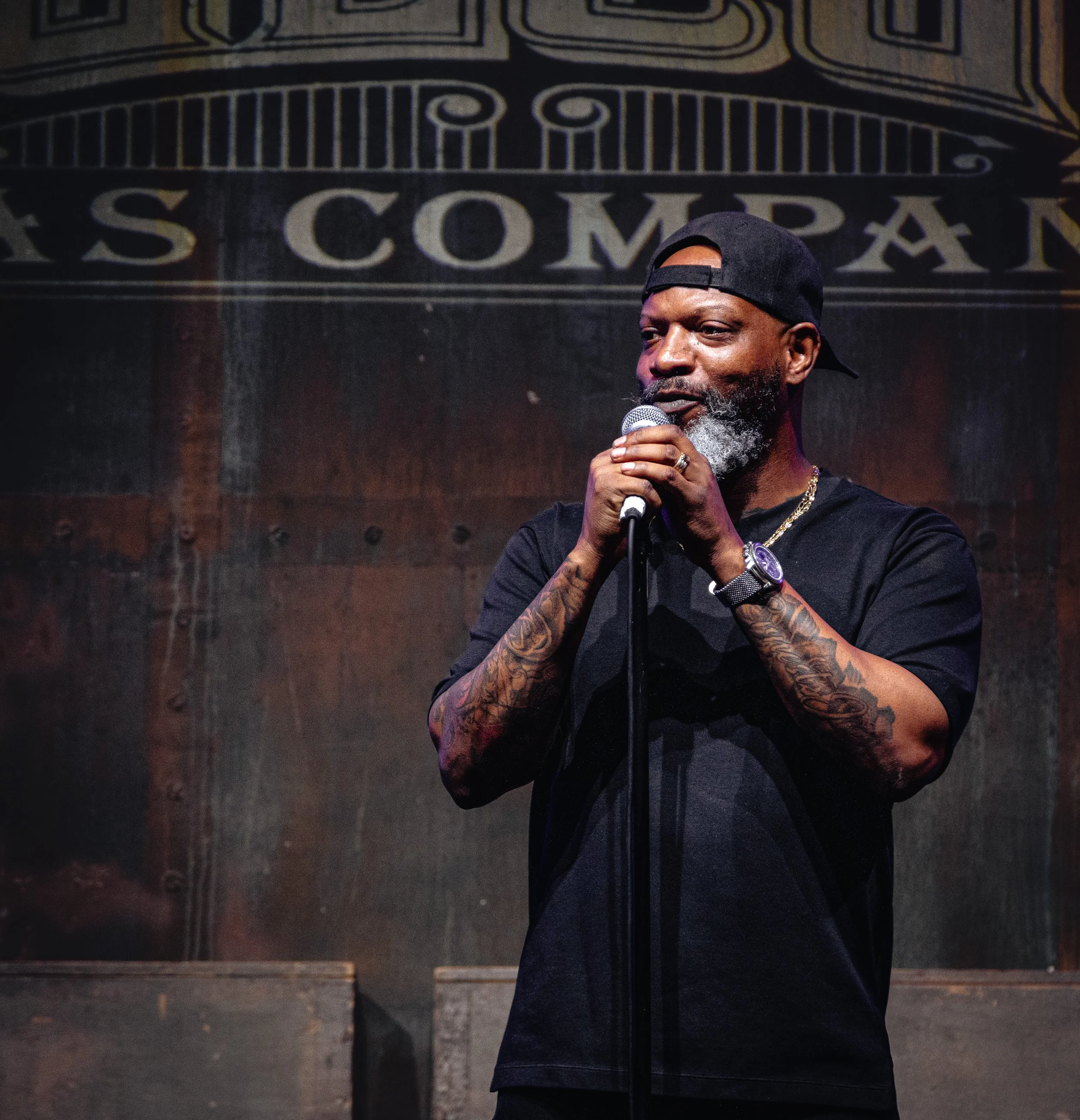 A man with a beard and tattoos, wearing a black baseball cap backward, holding a microphone, standing in front of a dark wall with a large sign that reads 'JS Company'.