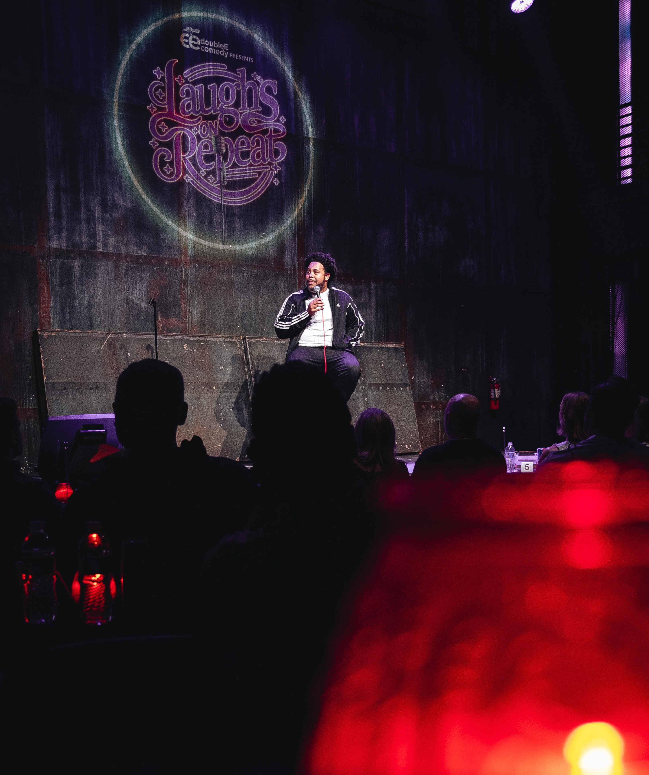 A performer sitting on stage with a microphone, under a screen displaying a sign that reads 'Laughs on Repeat', with audience members visible in the foreground.
