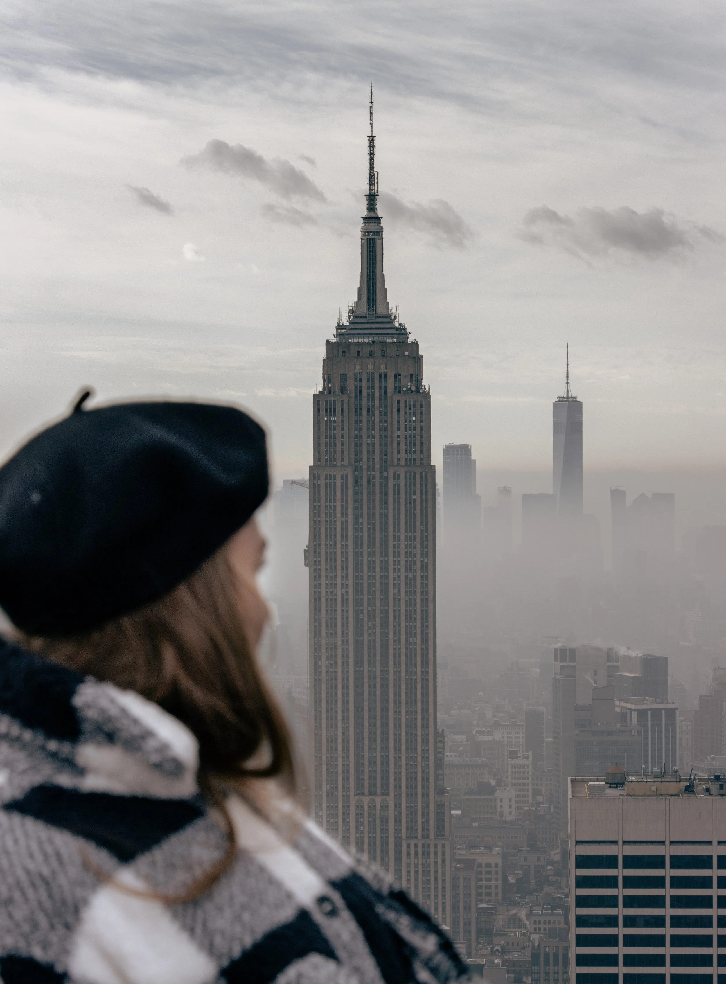 A person wearing a black beret and a black and white striped sweater looking at the Empire State Building in New York City, with another skyscraper visible in the background under a cloudy sky.