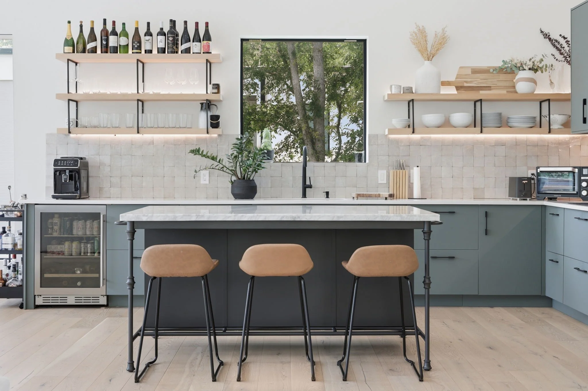 Modern kitchen with upper open shelves, a potted plant on the countertop, and a window showing green trees outside.