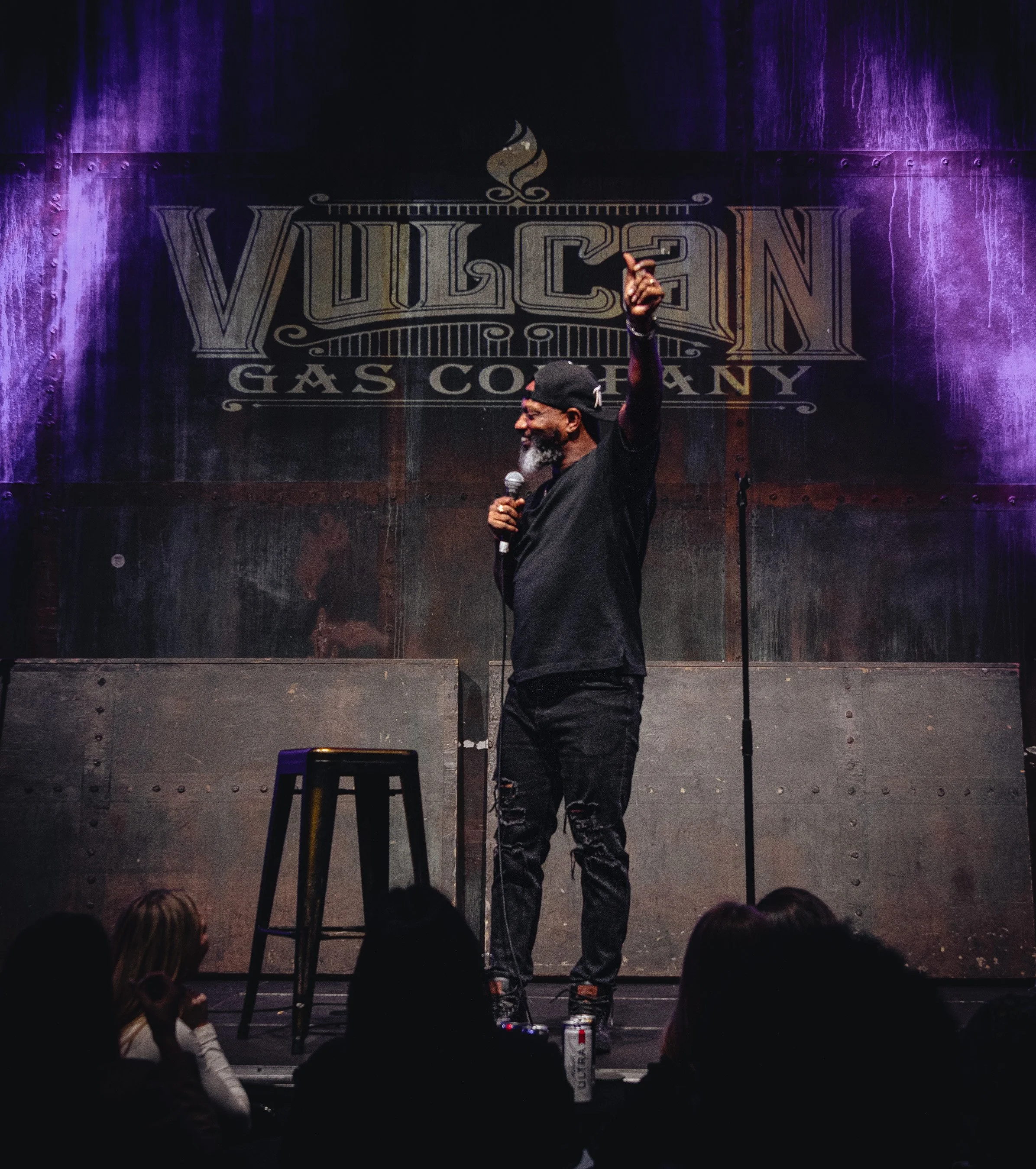 A man with a beard and black cap holding a microphone on stage, raising his right hand, with the word 'VULCAN' and 'GAS COMPANY' on the backdrop behind him. Audience members are visible in the foreground.