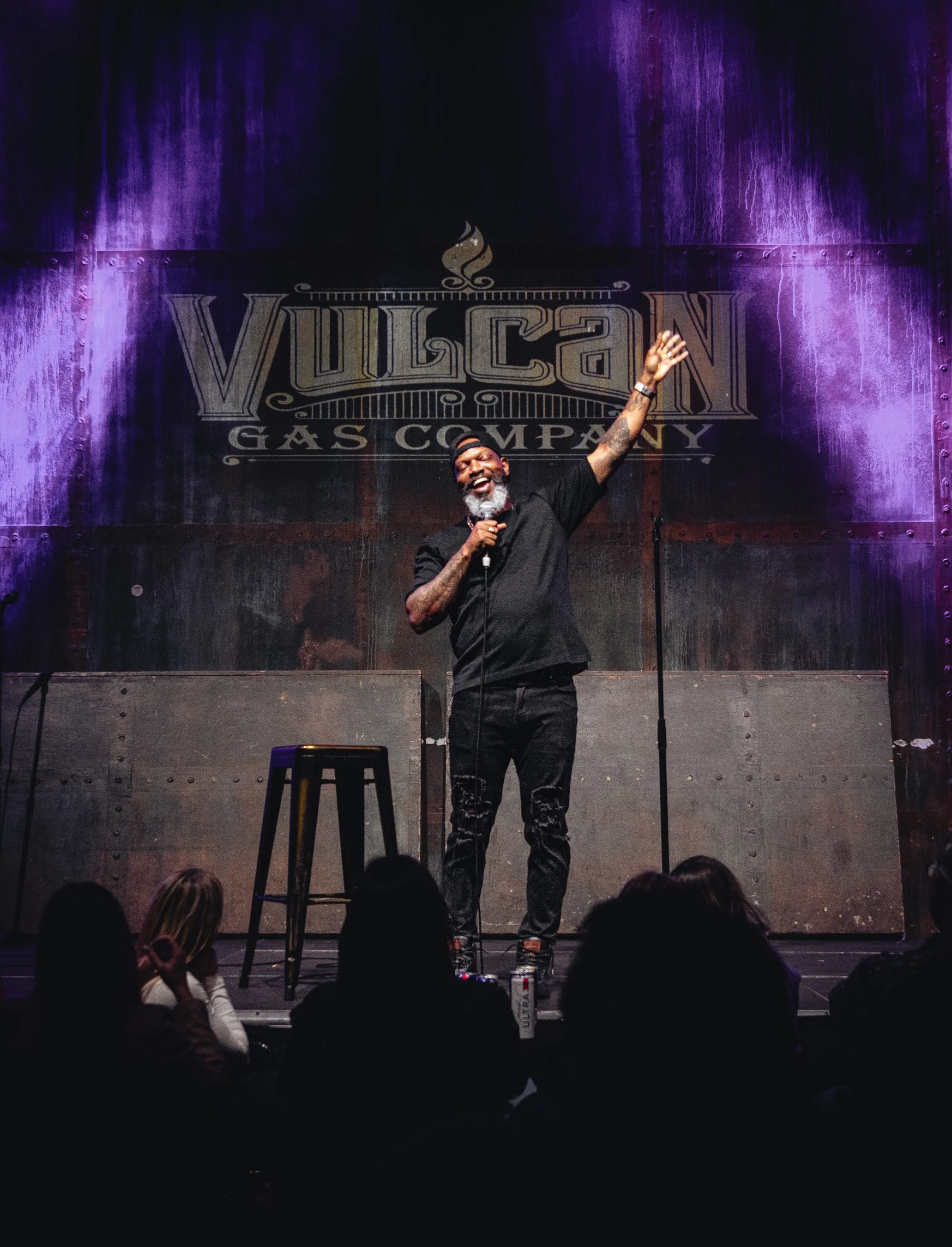 A man with a beard, wearing a black shirt, black pants, and a backwards black cap, is standing on a stage with a microphone in hand, smiling, and waving to an audience in front of a dark industrial-looking background with a sign that says 'Vulcan Gas
