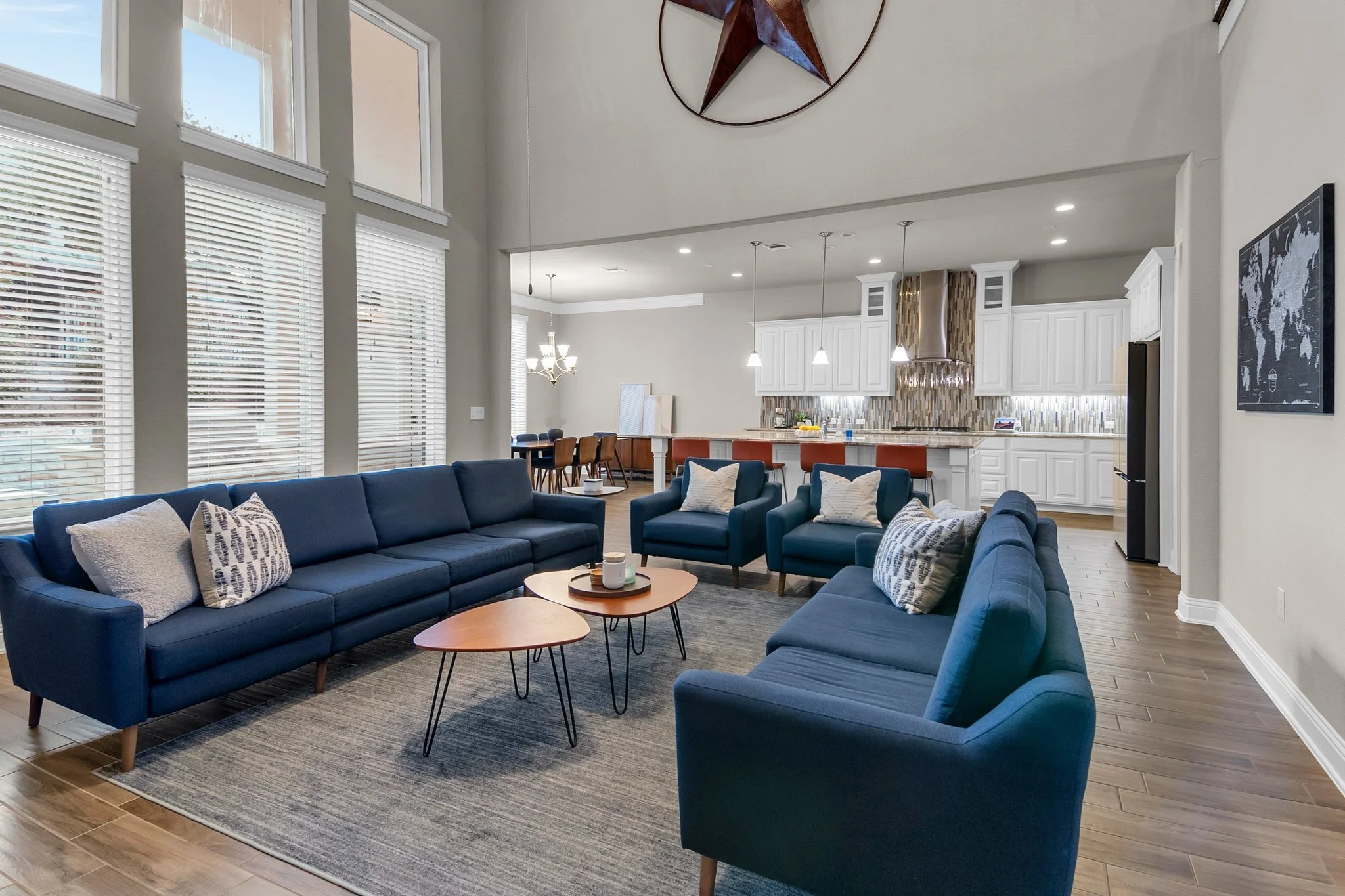Open-concept living room and kitchen with large windows, navy blue sofas, patterned pillows, wooden coffee tables, a blue area rug, white kitchen cabinets, a mosaic tile backsplash, and modern lighting fixtures.