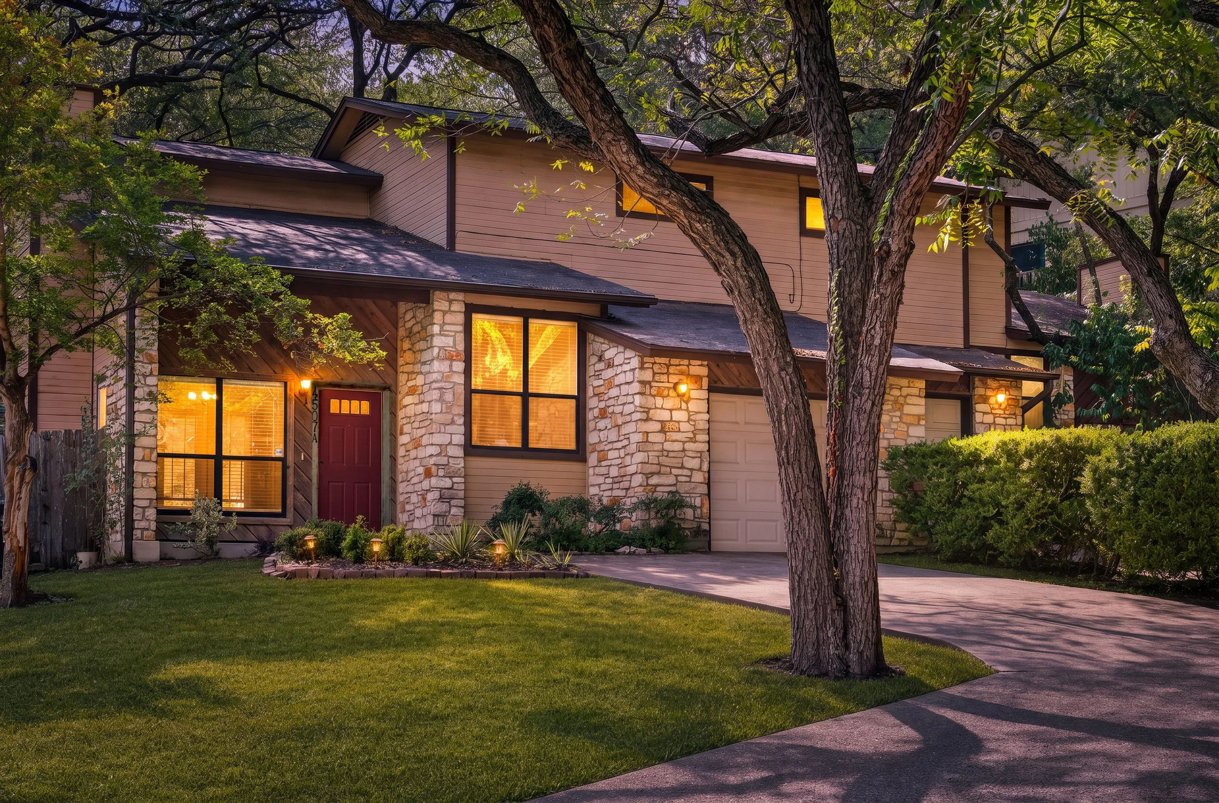 A two-story house with a stone facade, large windows glowing with warm light, surrounded by trees and a well-maintained lawn at dusk.