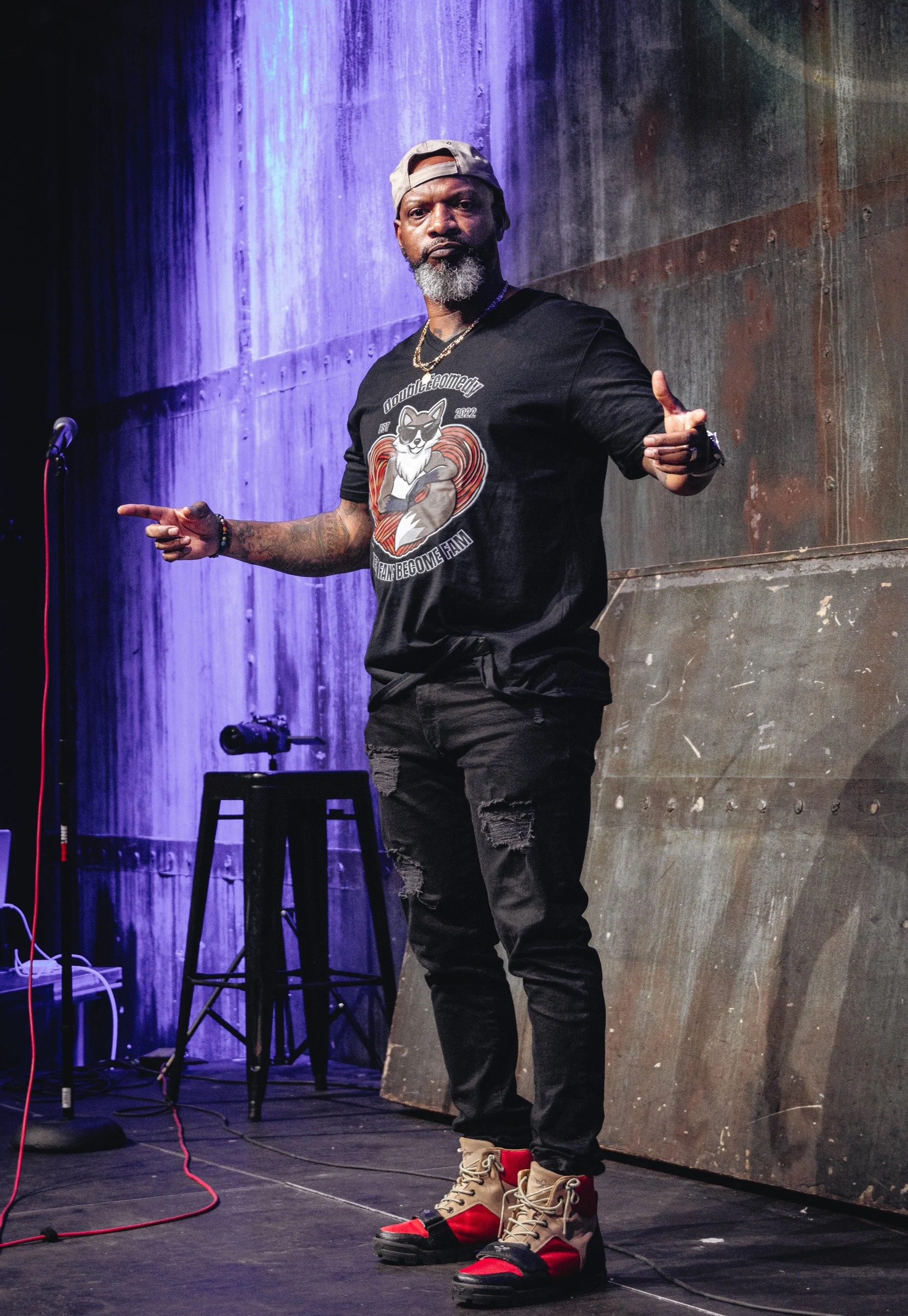 A man on stage in casual clothing, pointing with both hands. He wears a gray cap backwards, a black T-shirt with a cartoon dog and text, ripped black jeans, and unique red, beige, and black combat boots. The stage has a black stool, a microphone, and