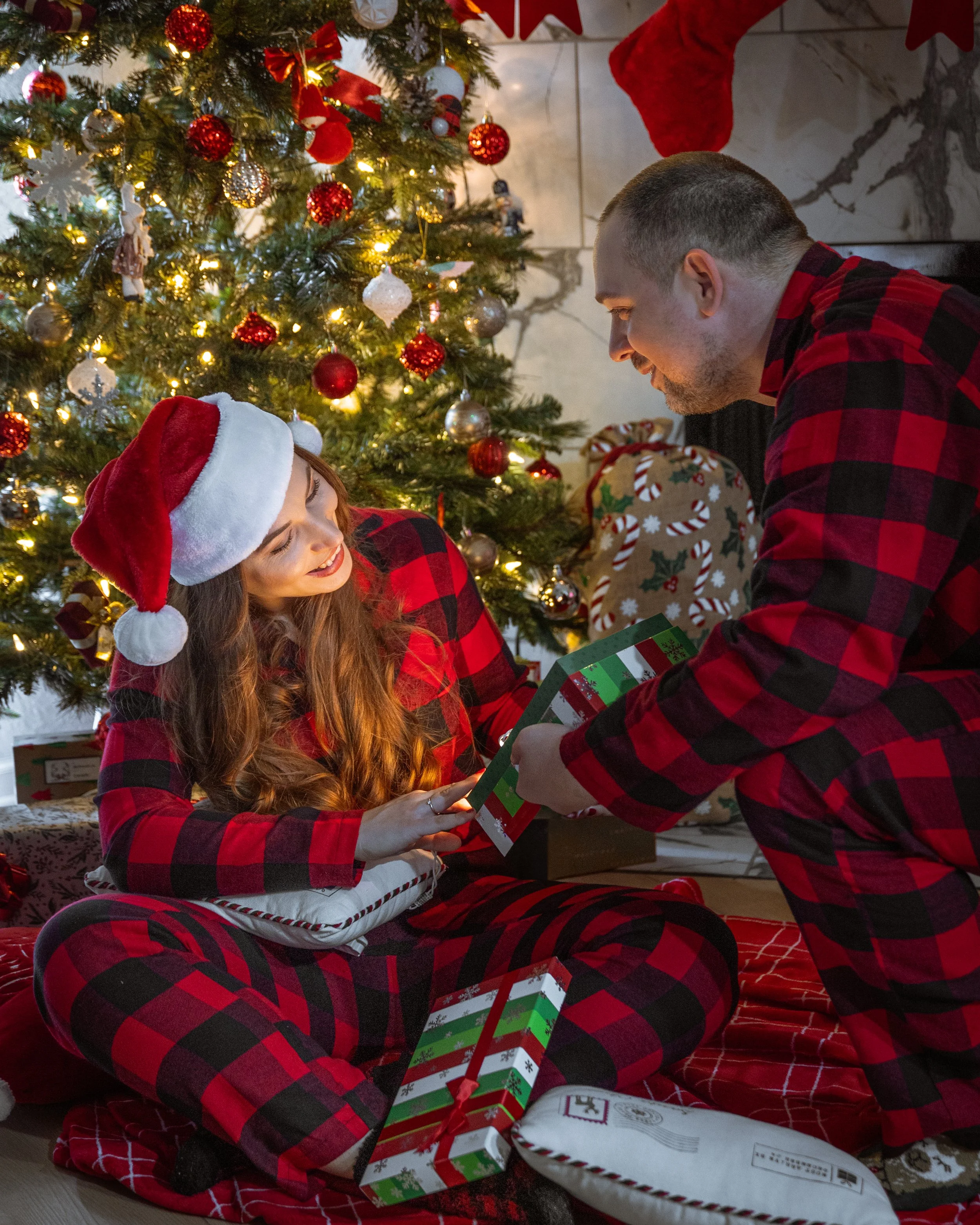 A man and woman in matching red and black plaid pajamas exchanging a Christmas gift in front of a decorated Christmas tree with lights and ornaments. The woman is wearing a Santa hat and sitting on the floor, while the man is kneeling. Several wrappe