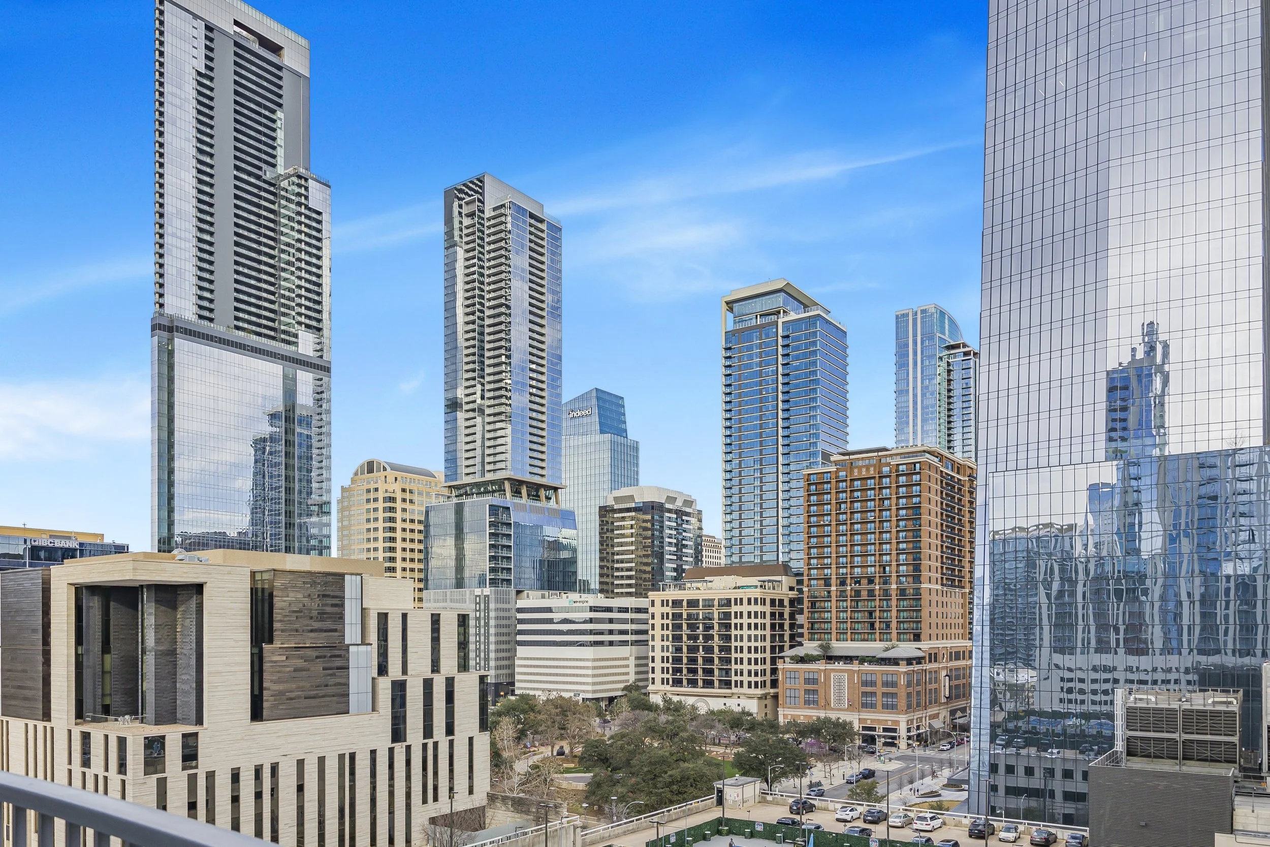 A city skyline with tall modern glass skyscrapers against a blue sky, with other mid-rise buildings and a park with trees at the bottom.