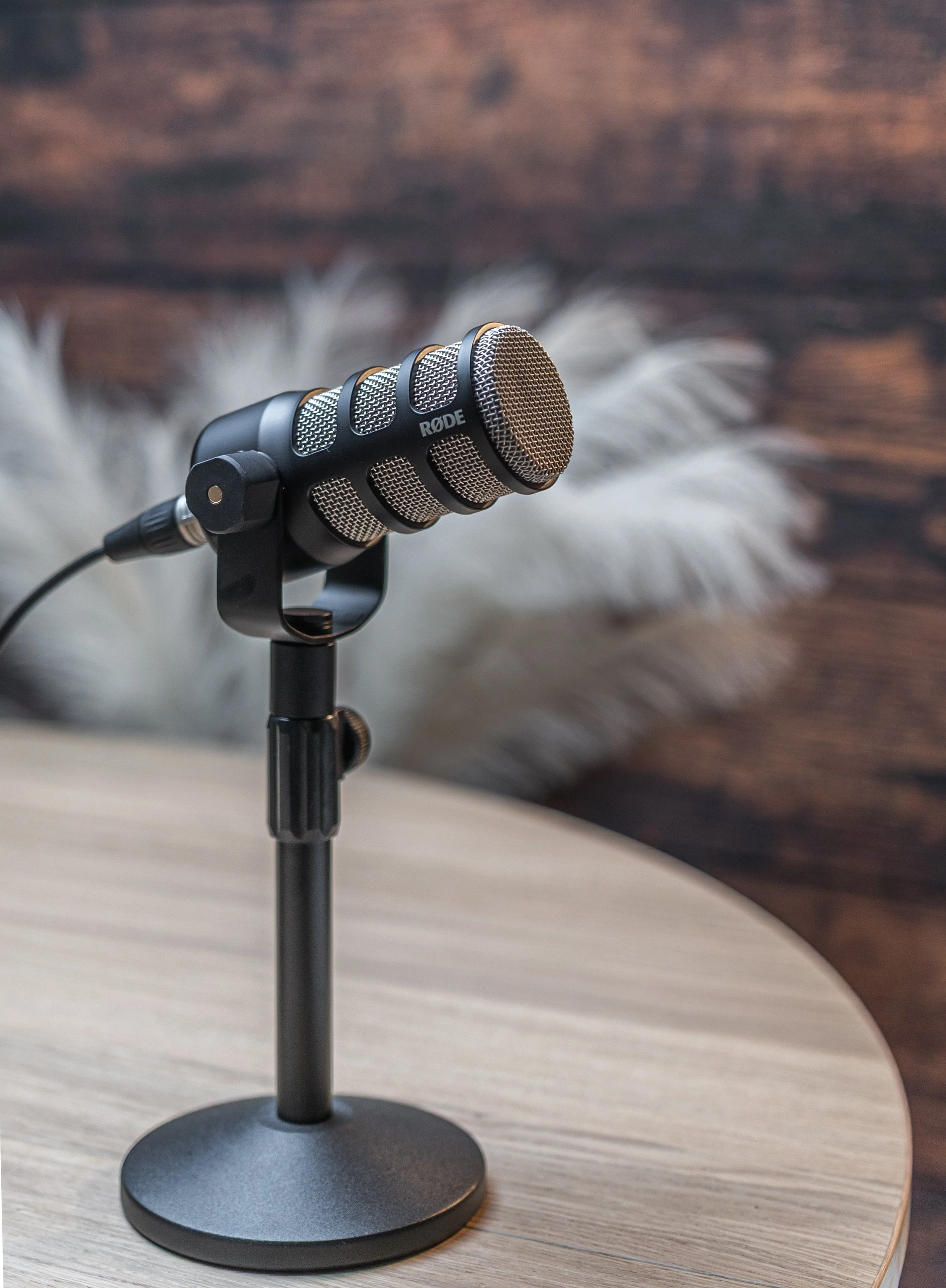 Close-up of a Rode microphone mounted on a stand, placed on a light-colored wooden table, with a blurred background.