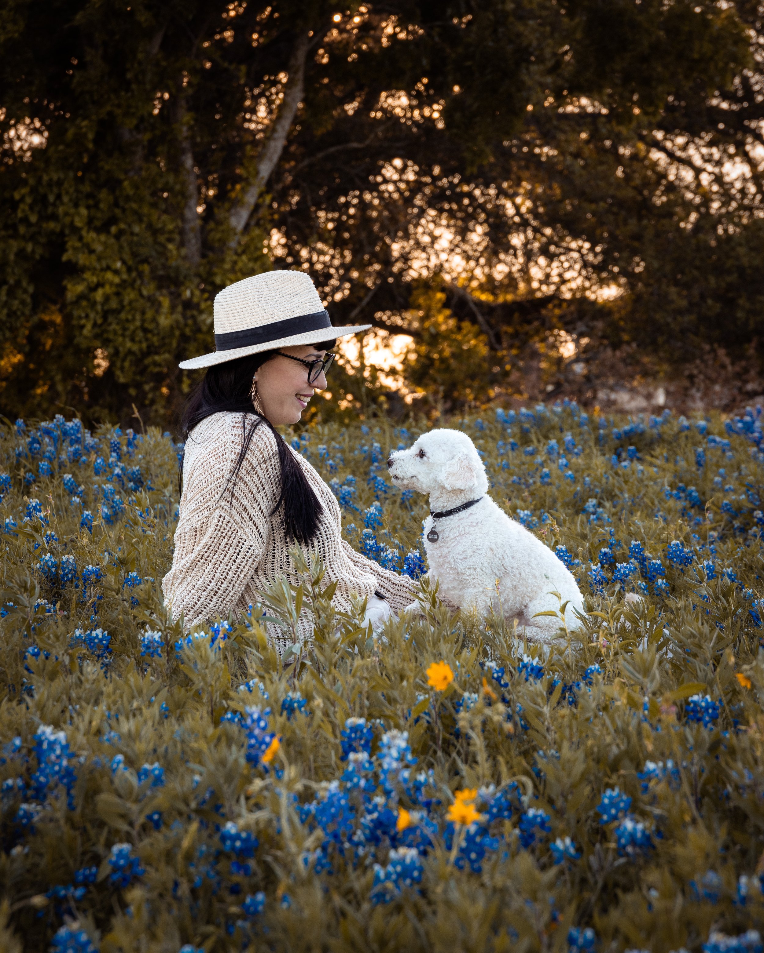A woman in a sweater and glasses wearing a wide-brimmed hat smiling while sitting in a field of blue and yellow flowers with a white dog, trees, and sunset in the background.