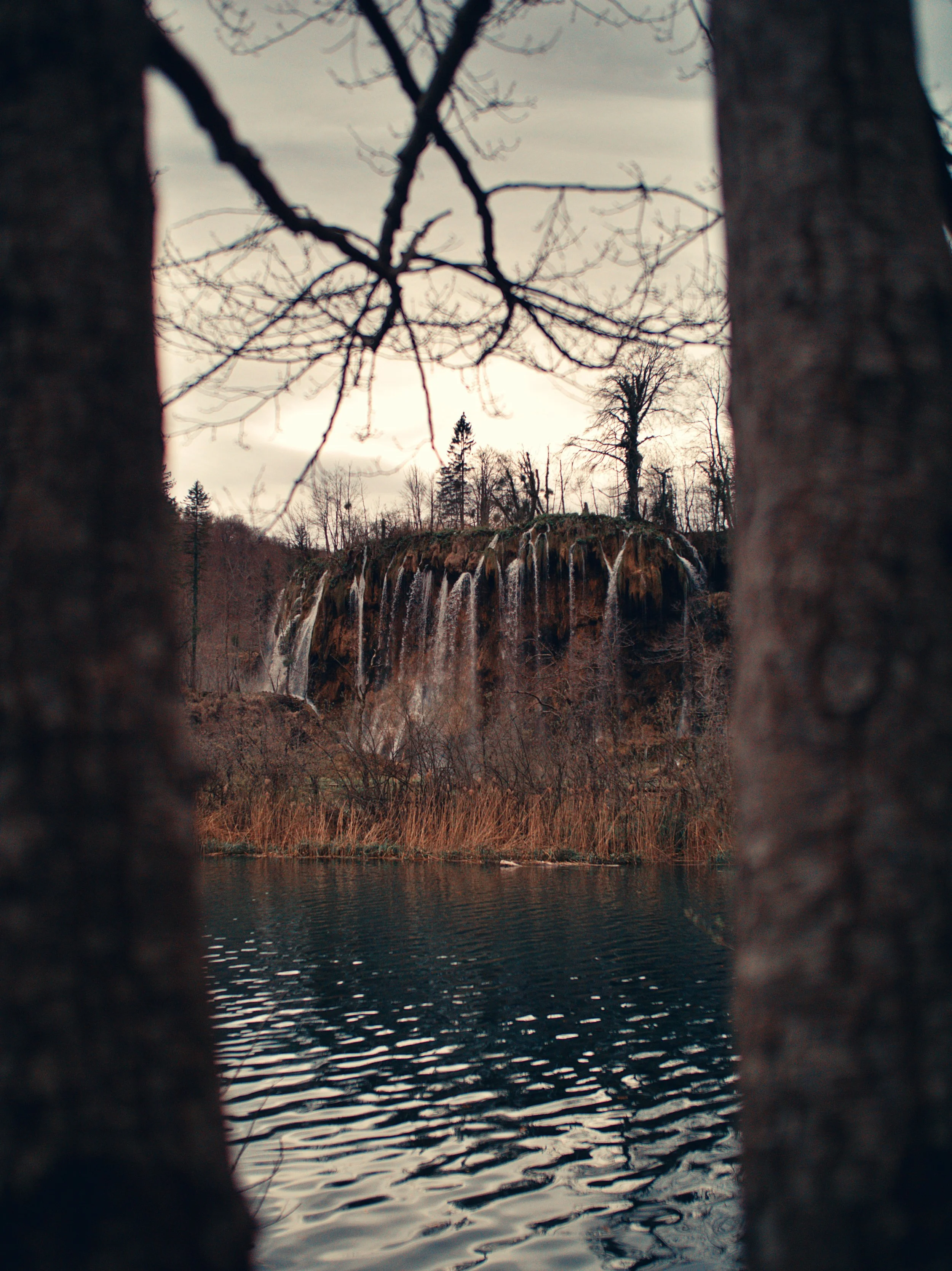 A view of a waterfall and trees seen through two gaps in a wooden fence near a body of water, with overcast sky.