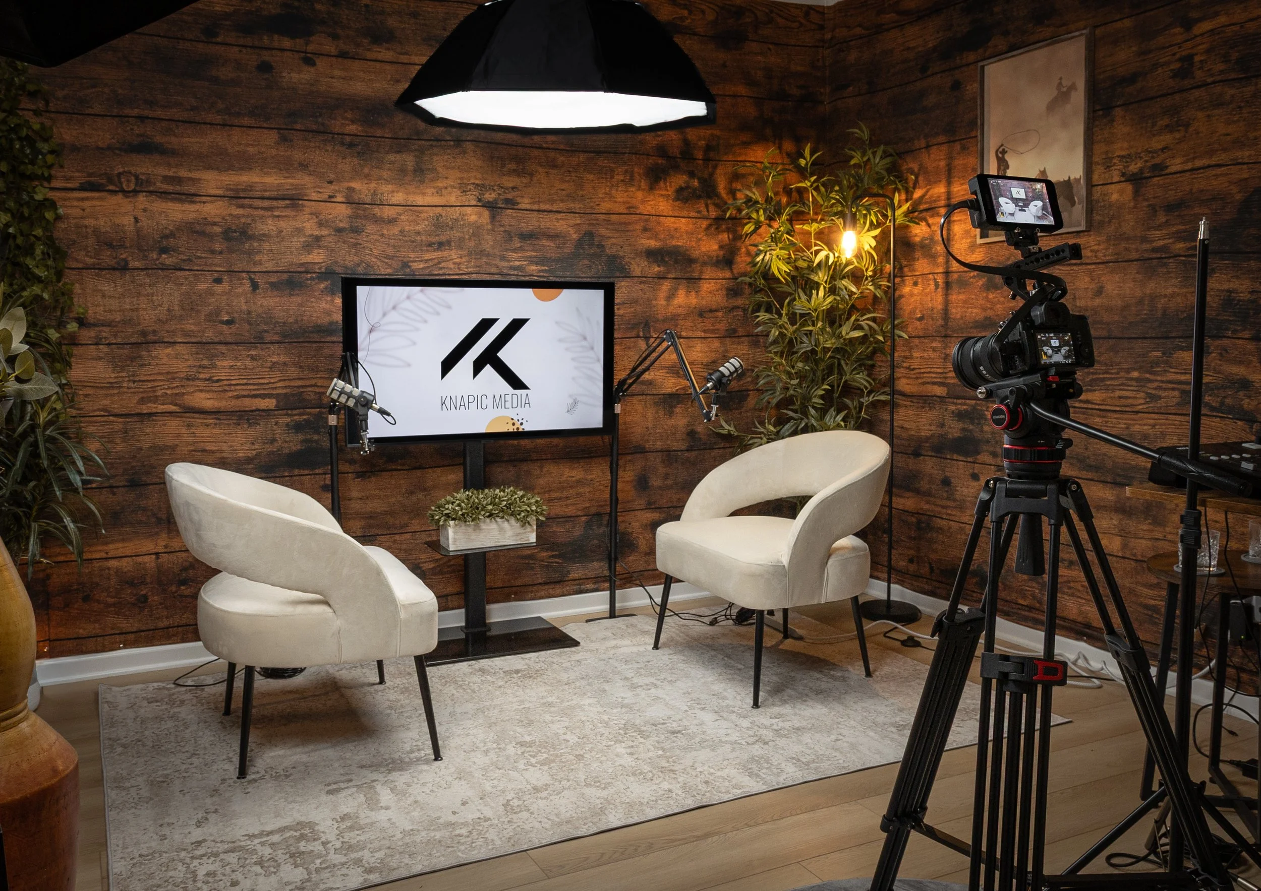 Setup for a video recording or interview in a room with wooden walls. There are two white armchairs facing each other, a small table with a plant, a television screen displaying the logo 'KNAPIC MEDIA,' professional camera equipment on a tripod, a la