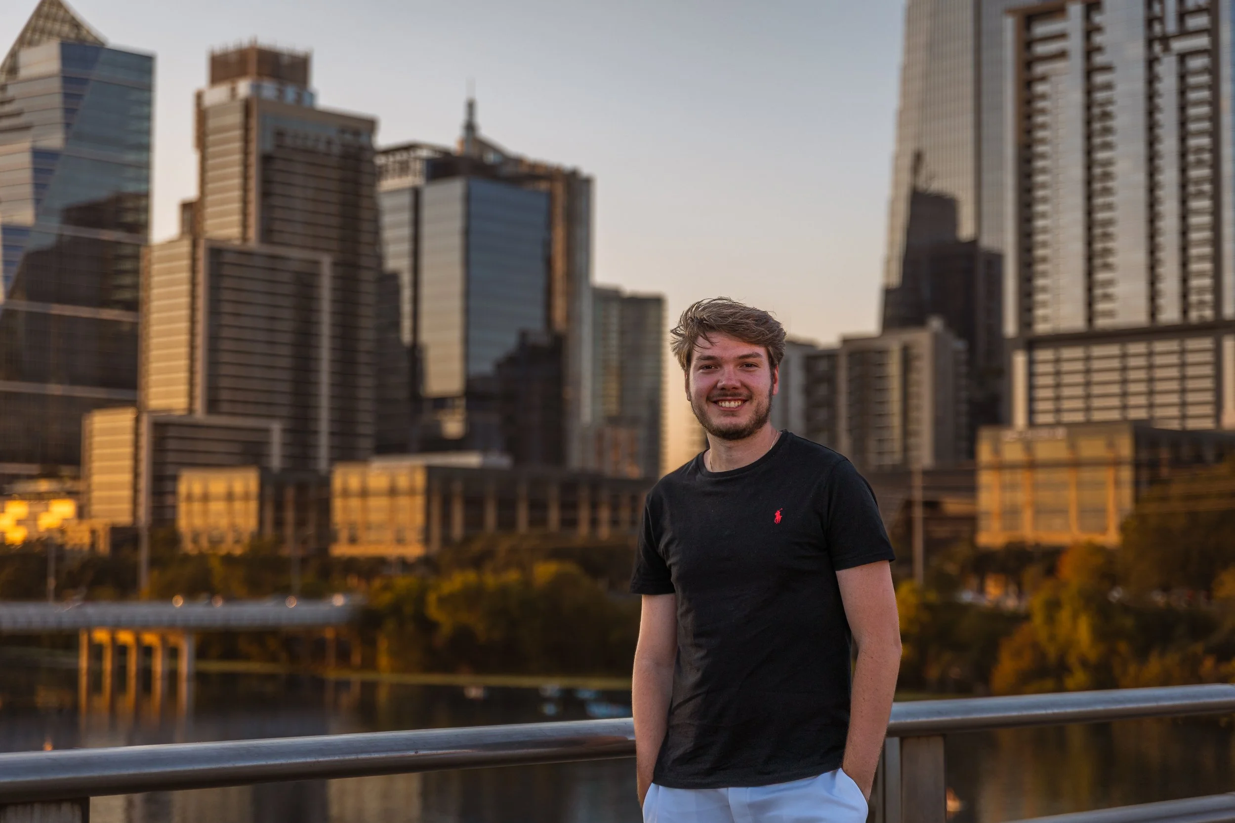A young man smiling outdoors on a bridge at sunset with a city skyline of tall buildings in the background.