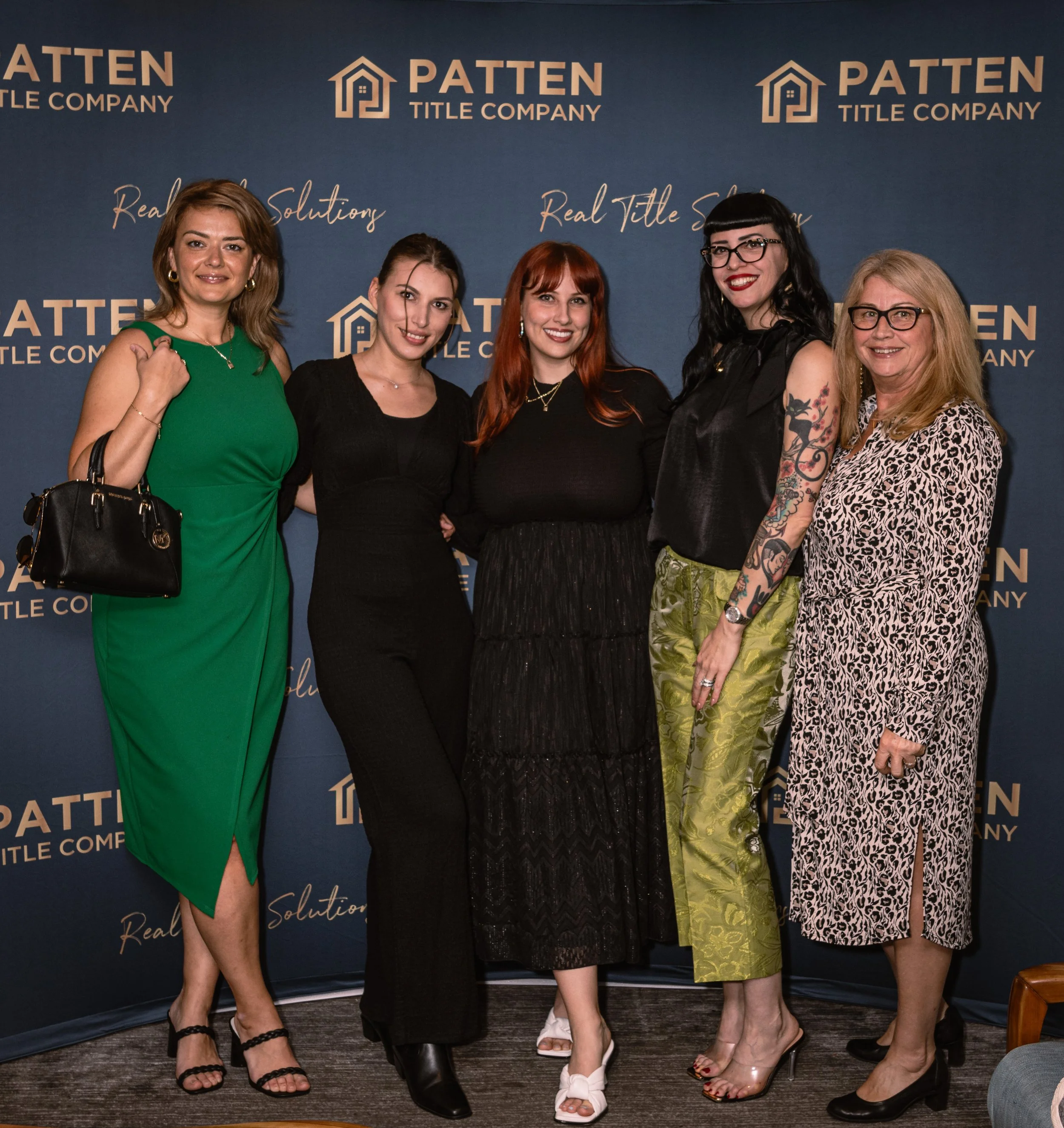 A group of five women standing in front of a blue banner with a logo and the words "PATTEN TITLE COMPANY". They are dressed in stylish clothing, smiling for the camera.