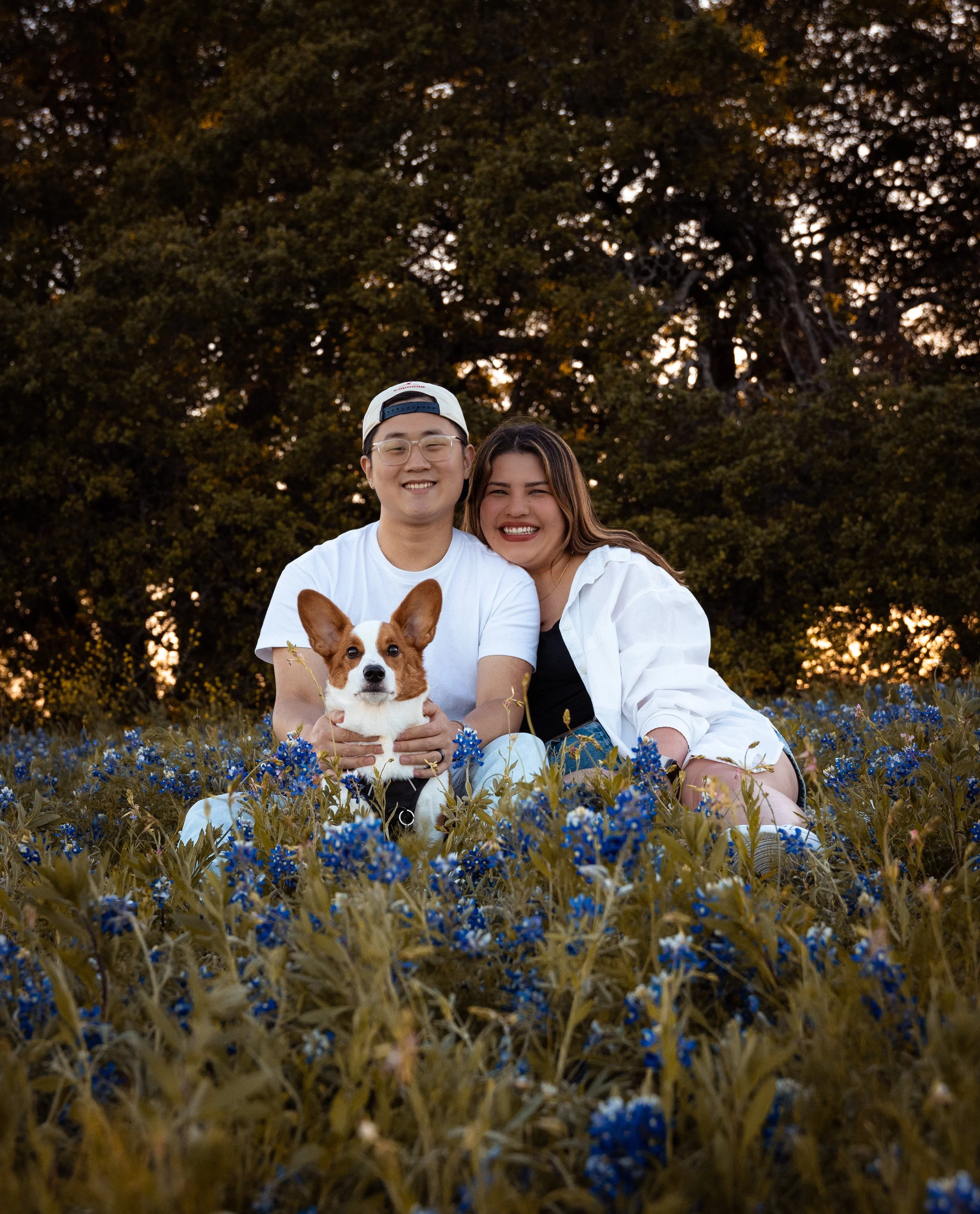 A young man and woman are sitting in a field of blue flowers, smiling at the camera. The man is wearing glasses, a white cap backwards, and a white shirt. The woman has long brown hair and is wearing a white shirt. A dog with large ears and a white and brown face is sitting in front of them. Behind them, trees are visible against the evening sky.