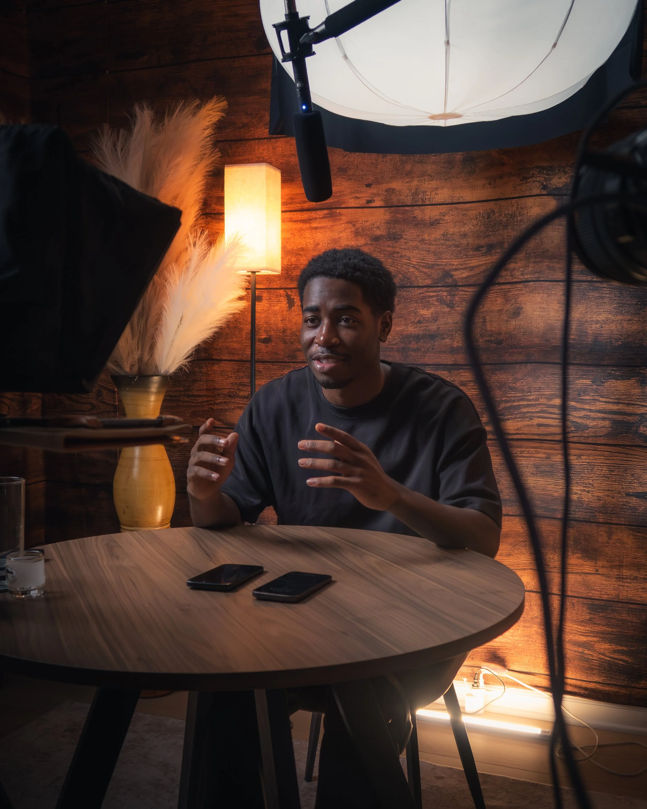 A man speaking during a video or photo shoot interview in a cozy room with a wooden wall, a lamp, and decorative feathers in a vase, with professional recording equipment visible.