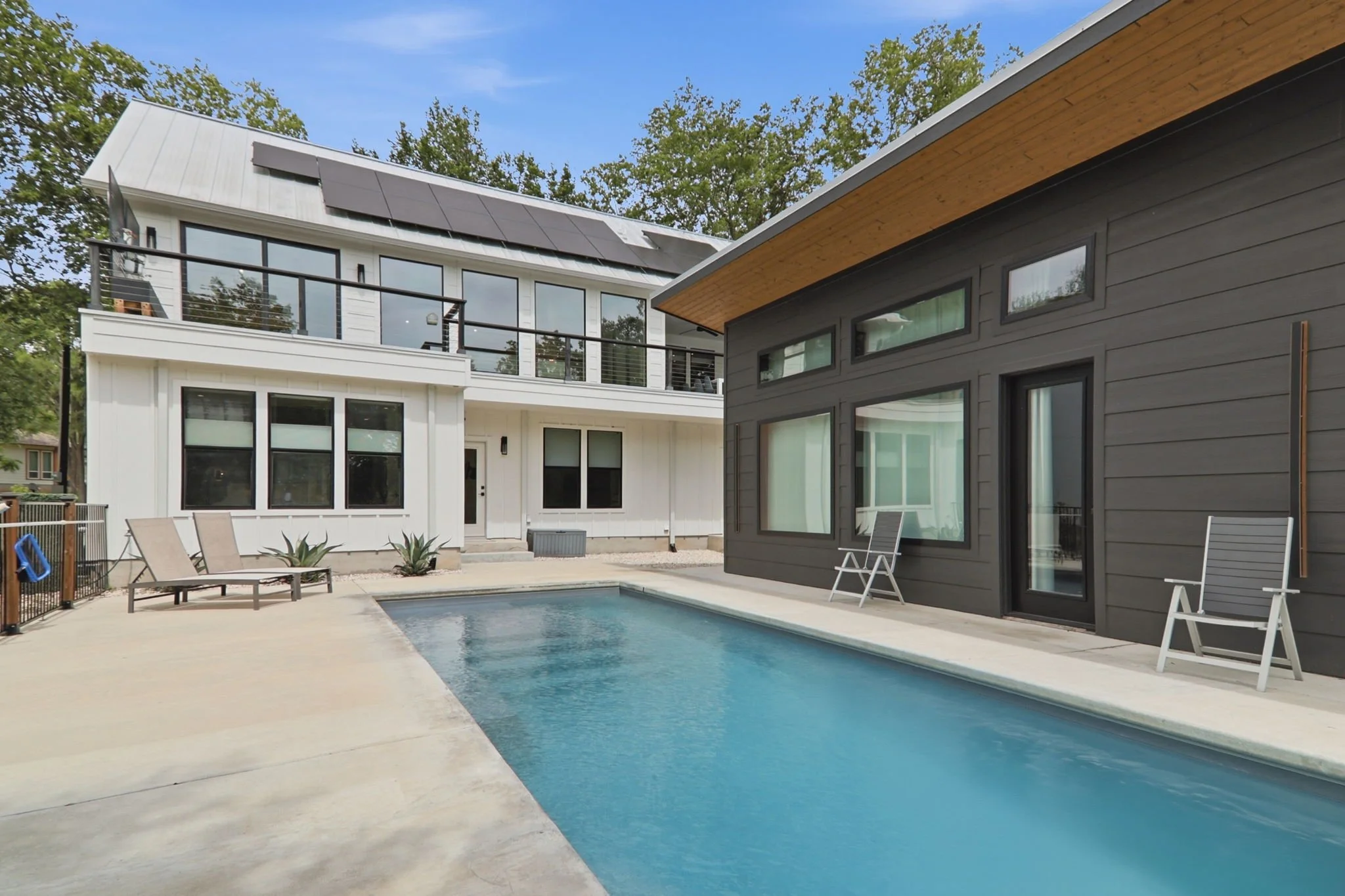 A modern backyard with a swimming pool, surrounded by a concrete deck, set against two adjacent houses with large windows and outdoor chairs, under a blue sky with trees in the background.