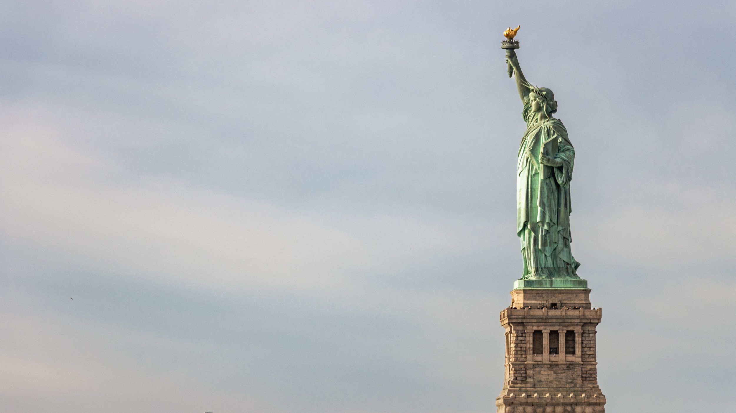 Statue of Liberty standing tall against a cloudy sky, holding a torch in her right hand and a tablet in her left, on a stone pedestal.