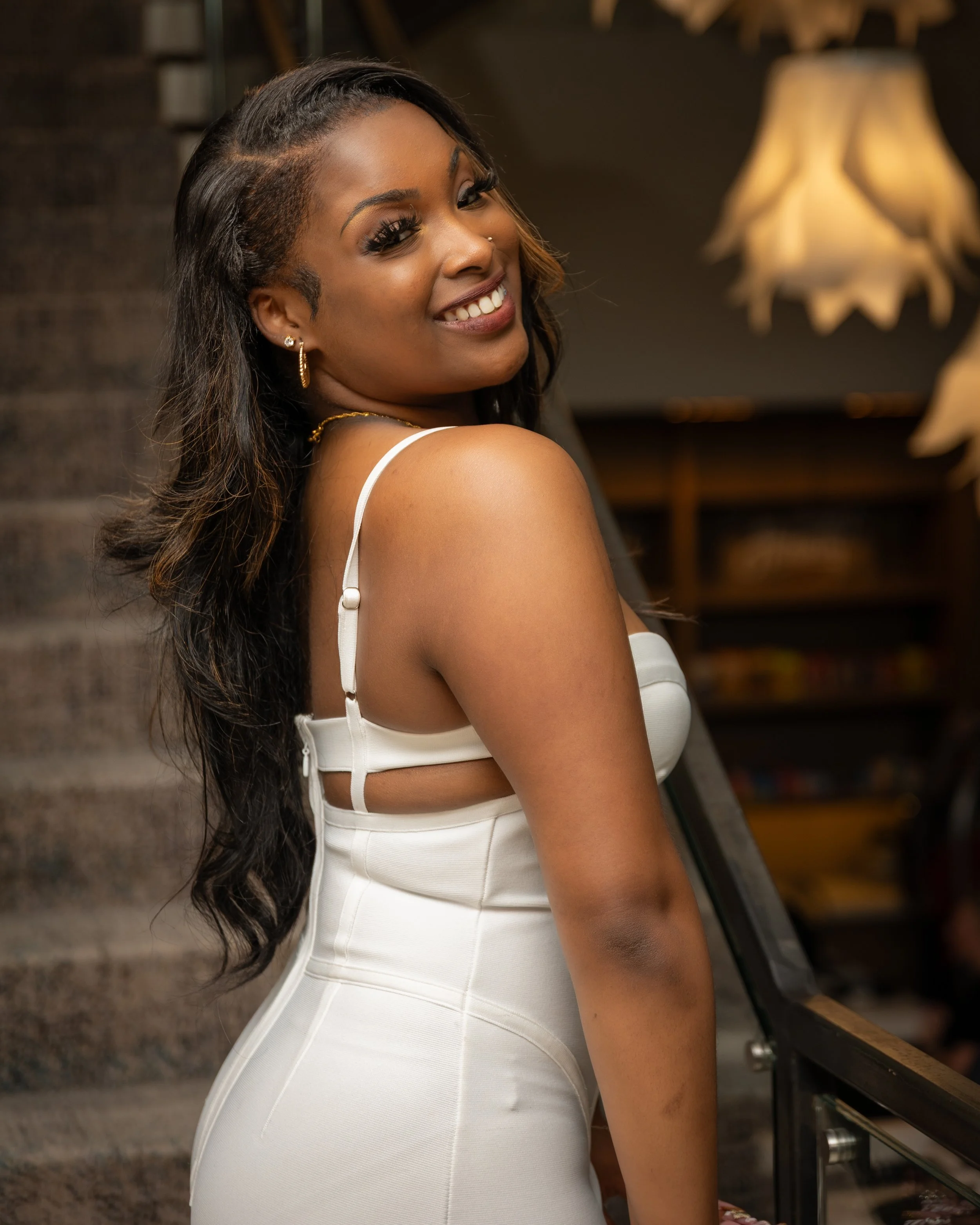 Close-up of a smiling woman with long dark hair, gold hoop earrings, and a small nose stud, wearing a white sleeveless dress, standing indoors with warm lighting and wooden shelves in the background.