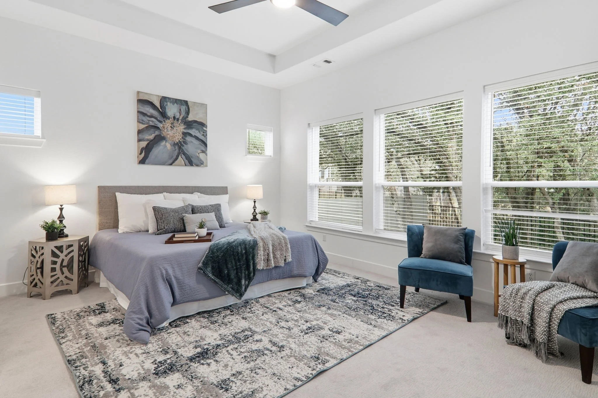 A bright, modern bedroom with white walls and large windows covered by white blinds. The bed has a grey upholstered headboard, white bedding, and decorative pillows. There are two matching nightstands with lamps on either side of the bed, and a small