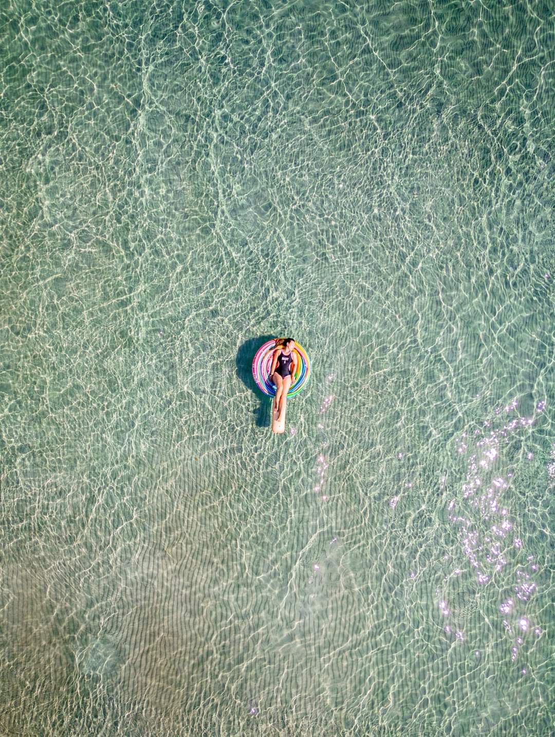 A person floats on a colorful inflatable ring in clear shallow water at the beach.