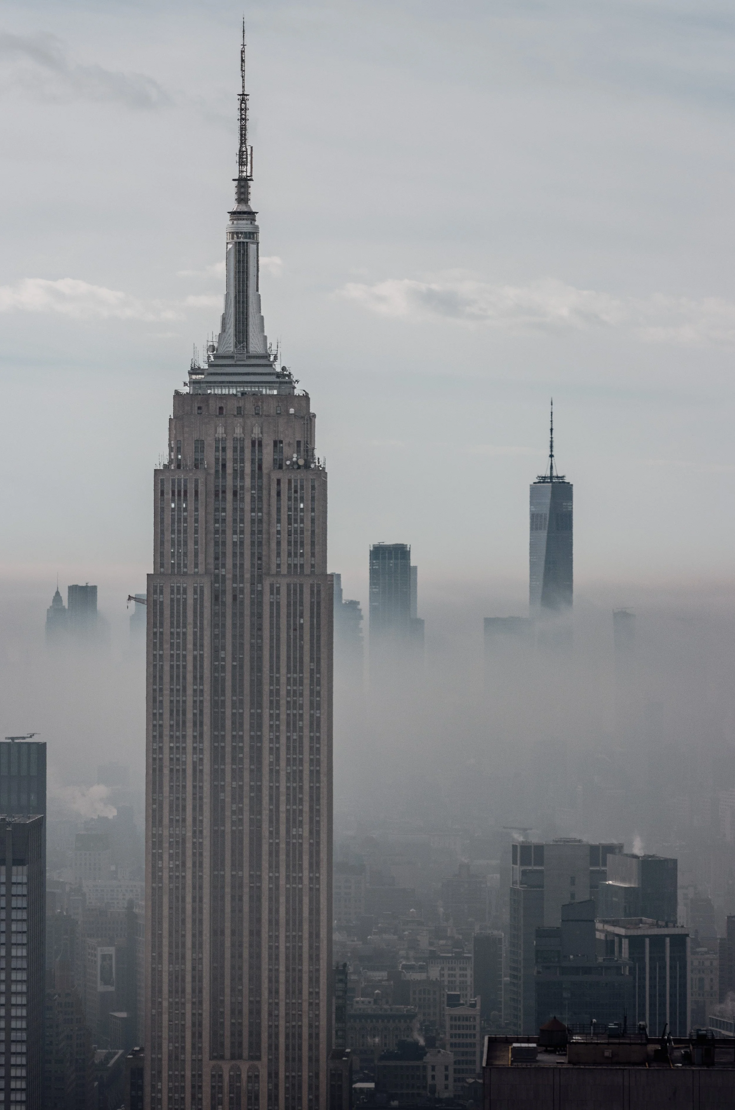 Skyline view of New York City with the Empire State Building and One World Trade Center visible, shrouded in fog and clouds.