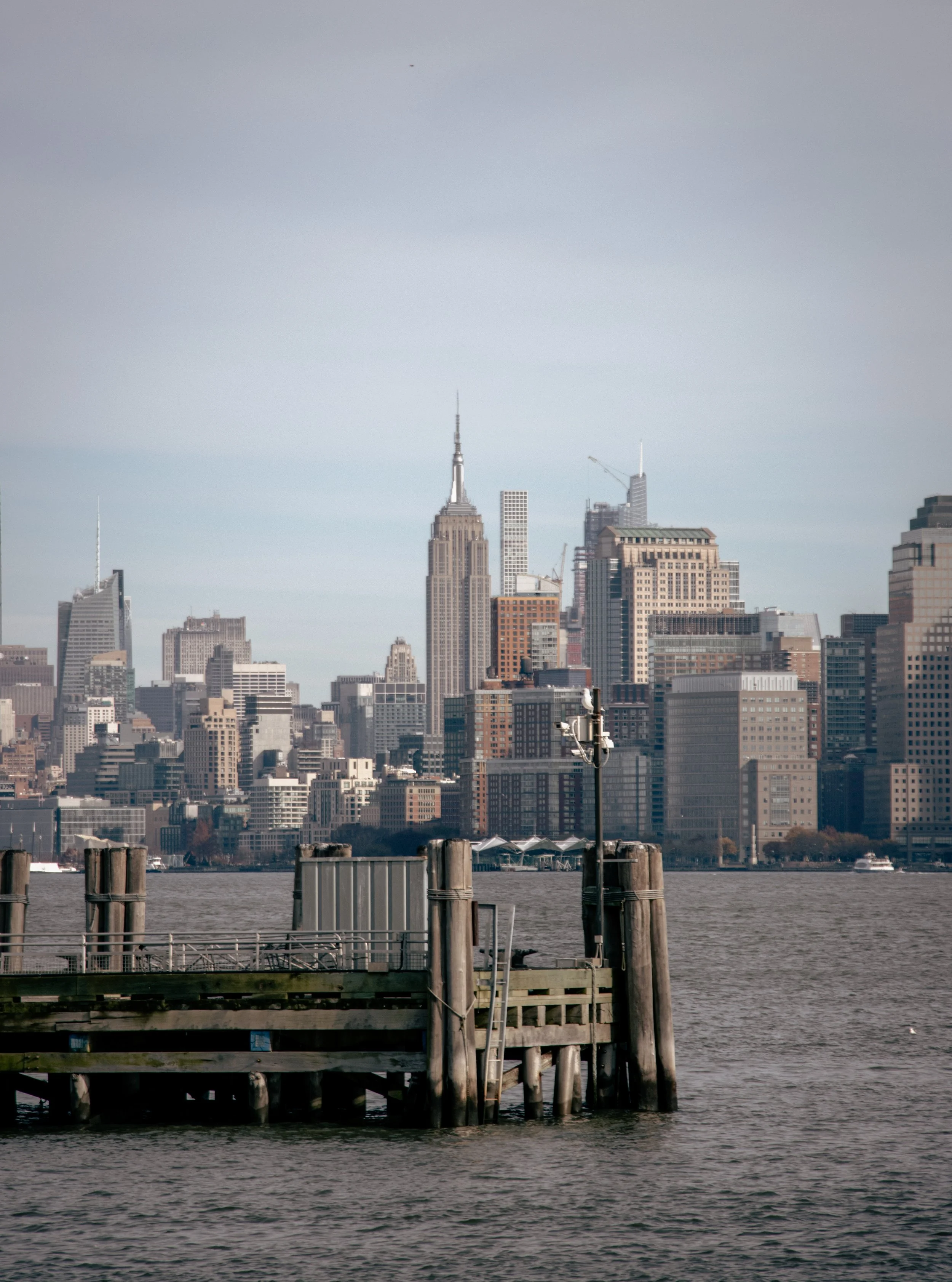 View of New York City skyline with buildings, including the Empire State Building, seen from the waterfront with wooden docks in the foreground and a boat on the water.