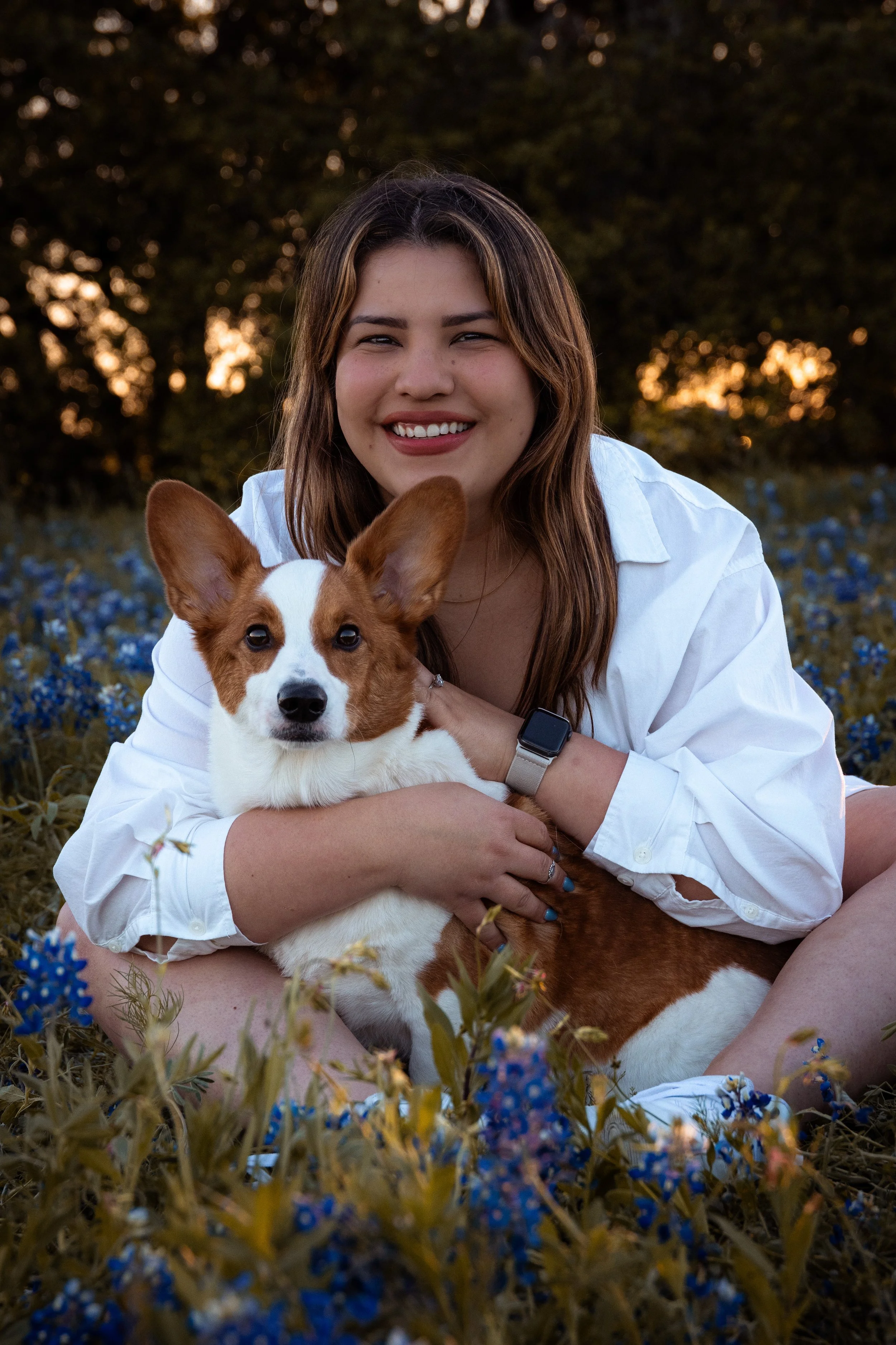 A woman with long brown hair, smiling, wearing a white shirt, sitting in a field of small blue flowers during sunset, holding a brown and white dog.