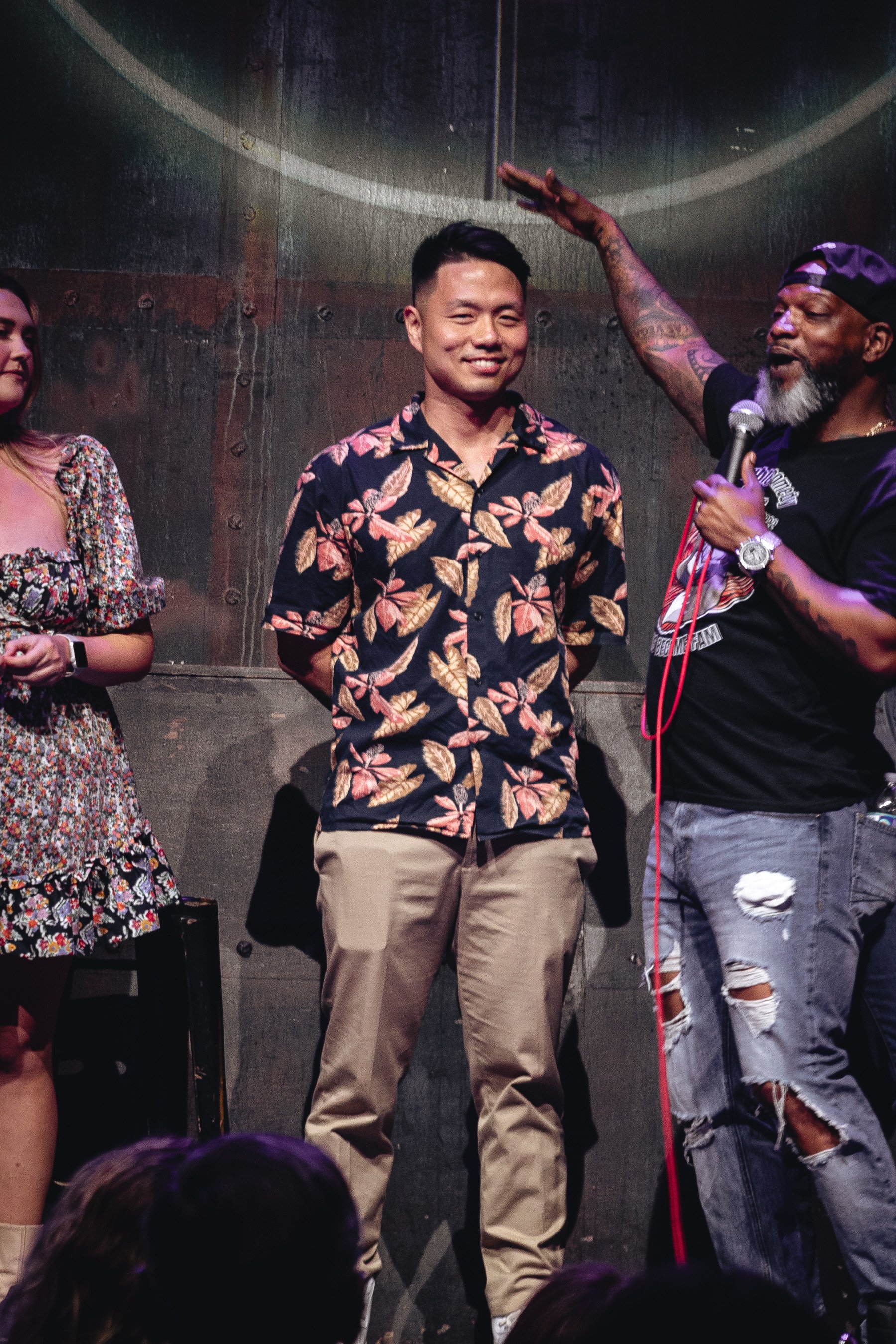 A young man in a floral shirt smiling at a stage performance, a man with tattoos and a cap holding a microphone gestures above him, and a woman in a floral dress is partially visible on the left, all against a dark industrial background.