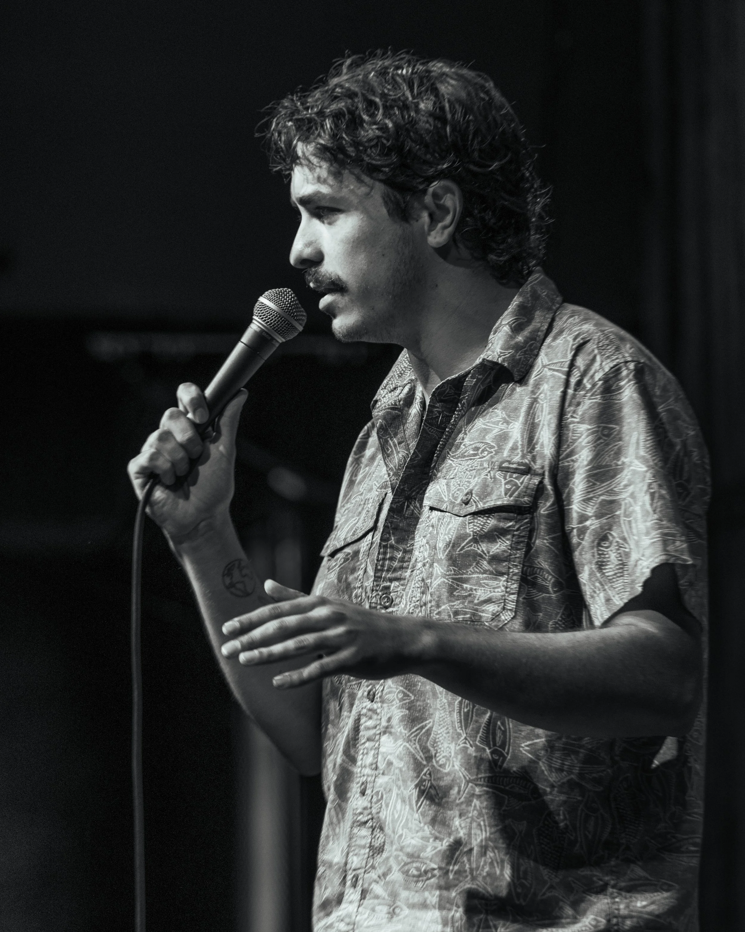 A black-and-white image of a man with curly hair and a mustache, speaking into a microphone. He is wearing a patterned shirt and is standing against a dark background.
