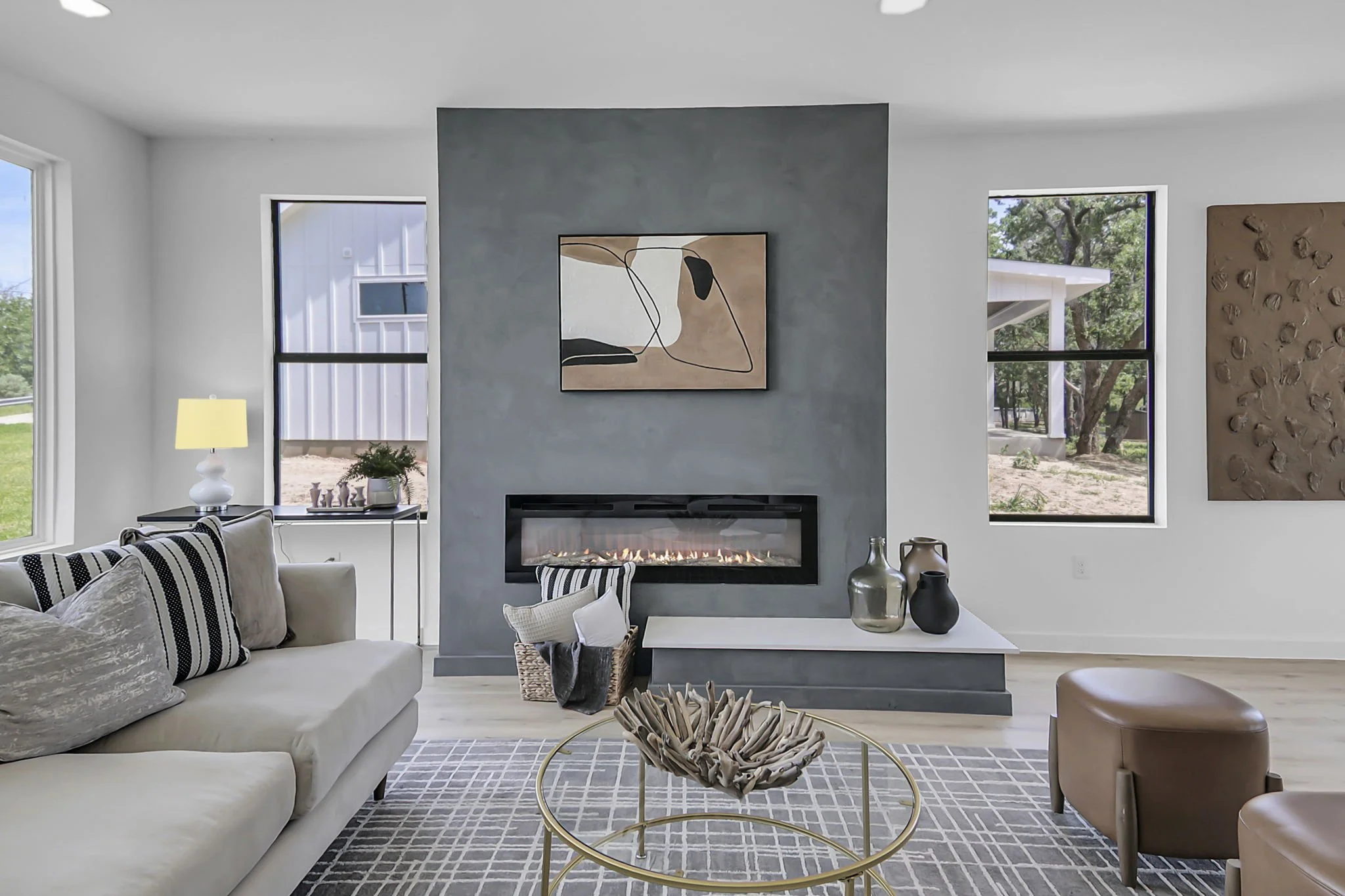 Living room with a gray accent wall, abstract art, fireplace, white sofa with striped pillows, side table with a lamp, wicker basket, decorative vases, windows showing outdoor trees and house.