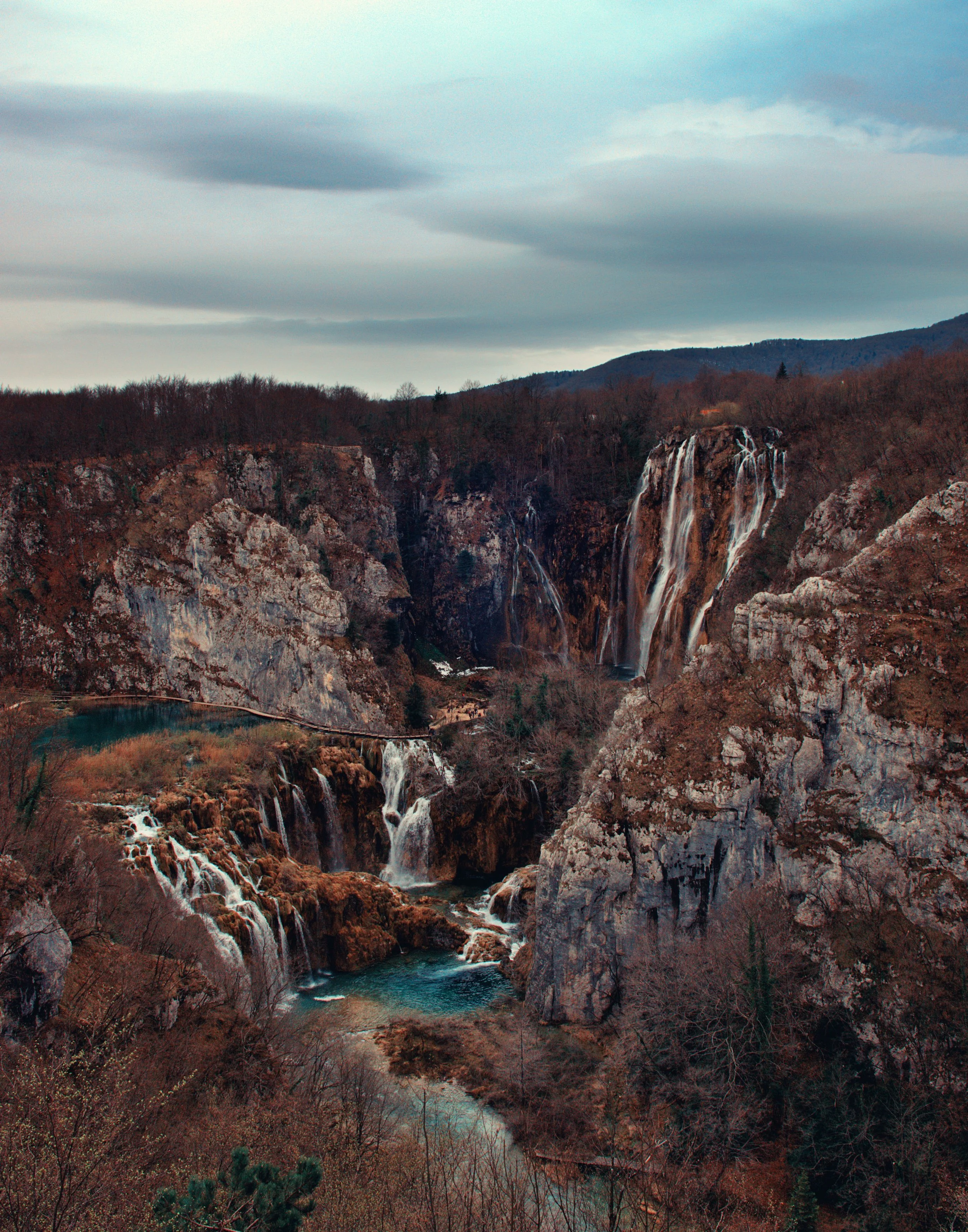 A scenic view of waterfalls on a rocky hillside with a forest in the background under a cloudy sky.