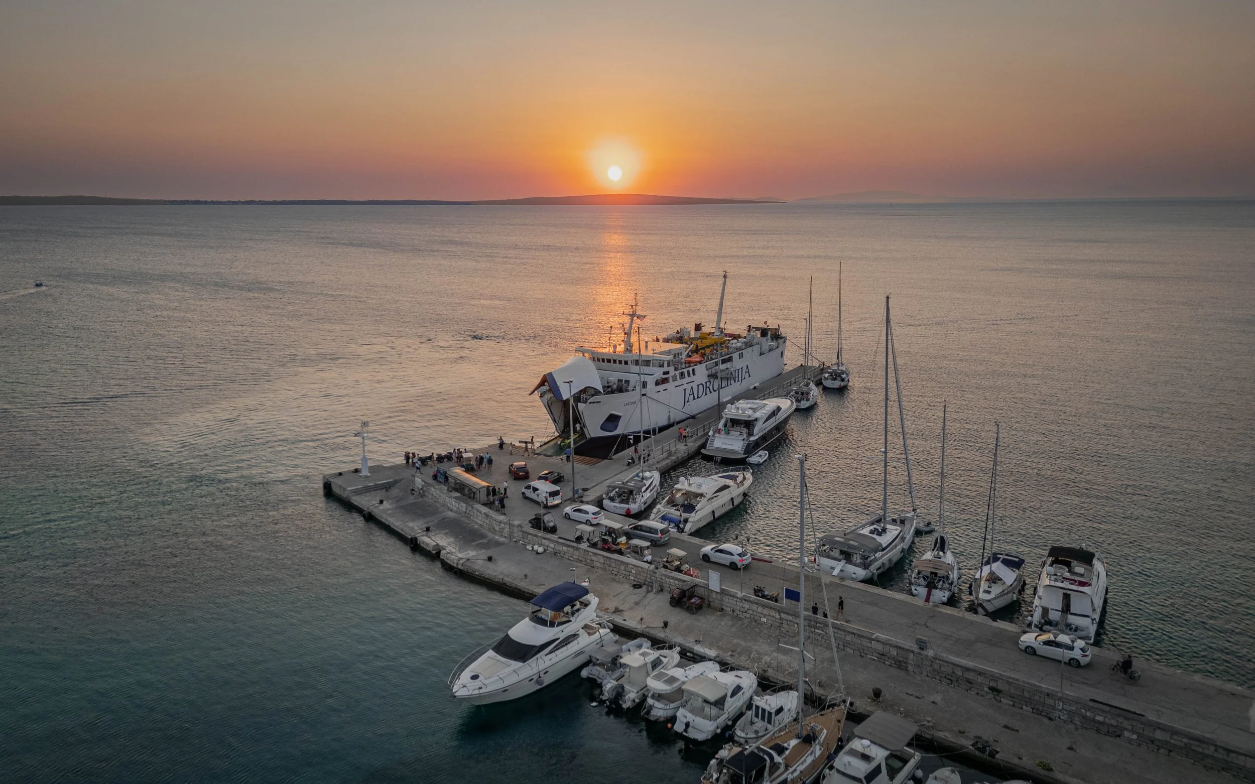 A harbor with boats docked along a pier during sunset, showing calm water and a distant horizon.
