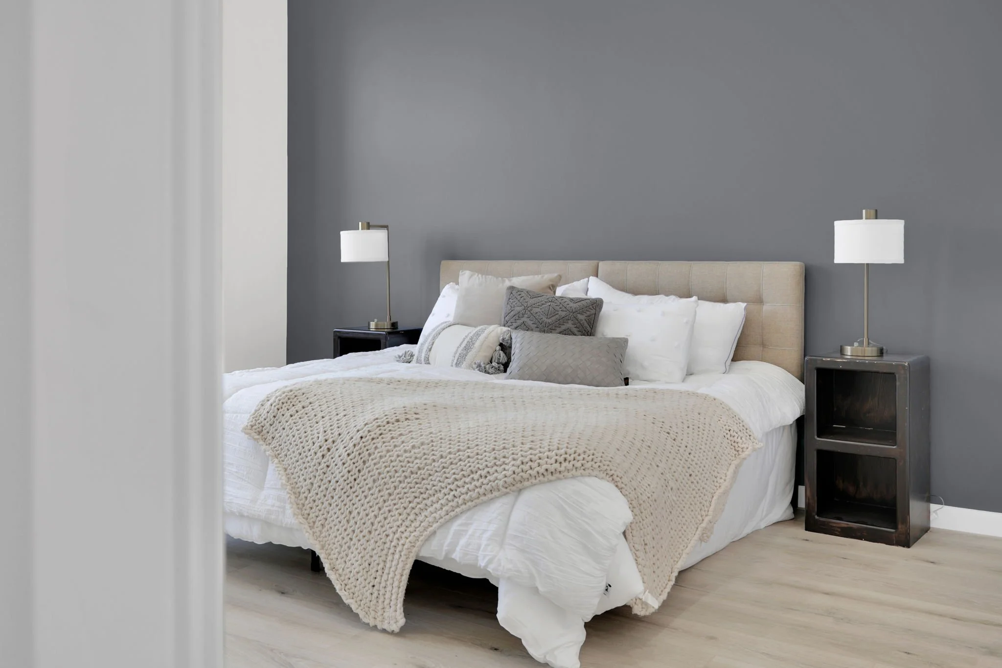 A neatly made bed with numerous pillows and a chunky beige blanket, flanked by two matching nightstands with lamps, in a modern bedroom with a gray accent wall and light wood flooring.
