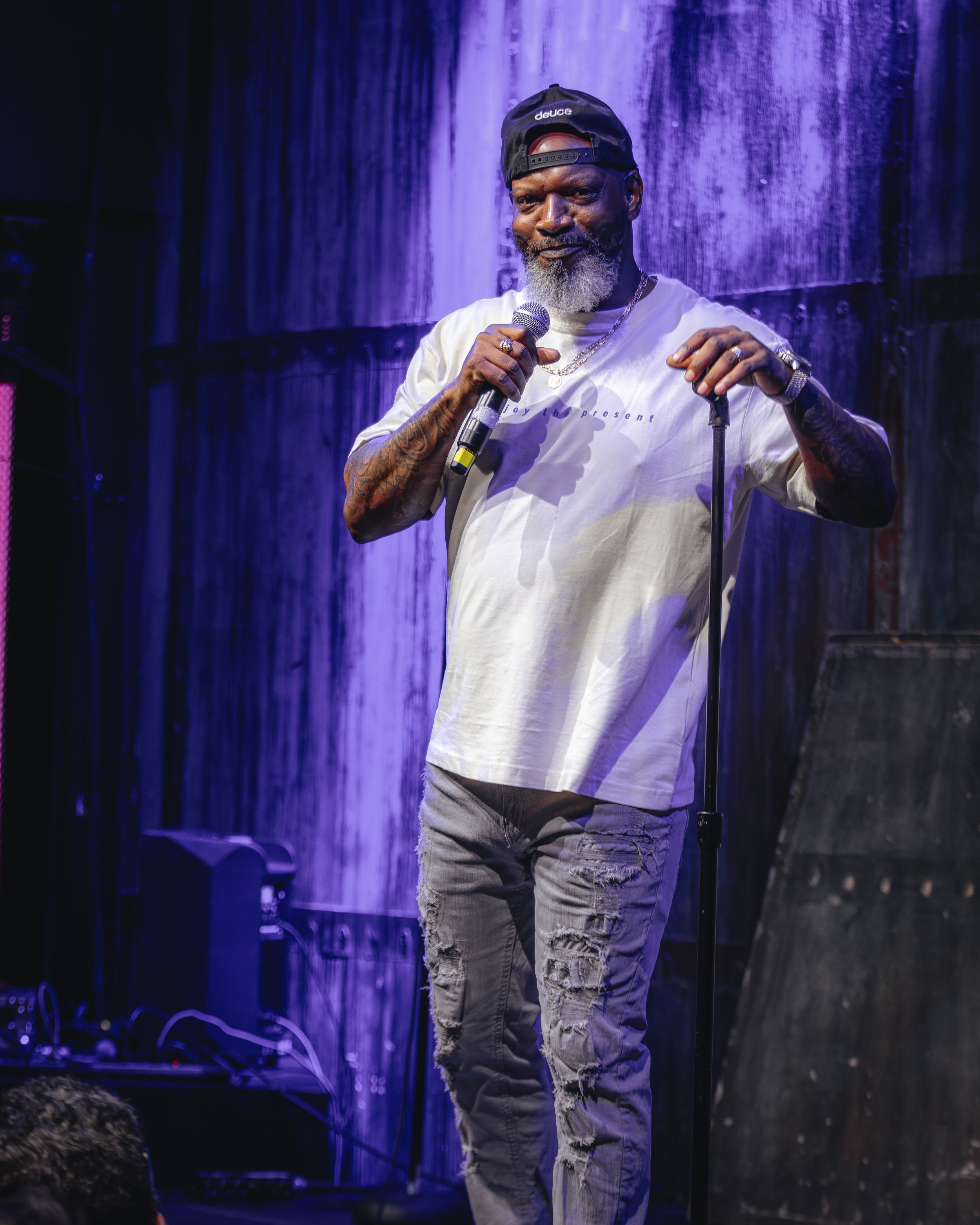A man with a beard, wearing a black backward cap, oversized white t-shirt, and ripped jeans, holding a microphone, standing on a stage with a waterfall backdrop, purple lighting illuminating the scene.