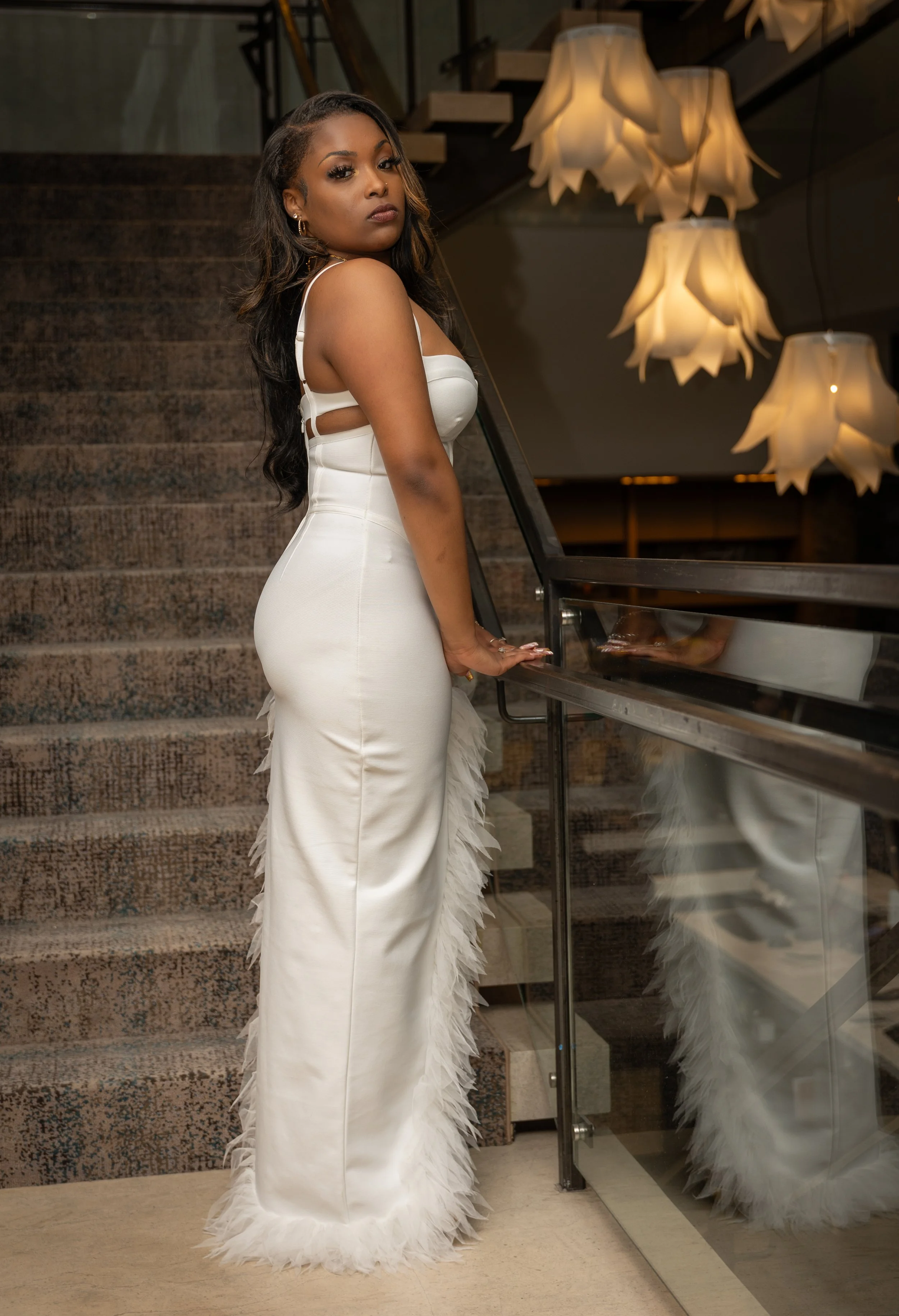 A woman wearing a white gown with feather details along the bottom, standing next to a mirrored wall and railing, with a staircase and pendant lights in the background.