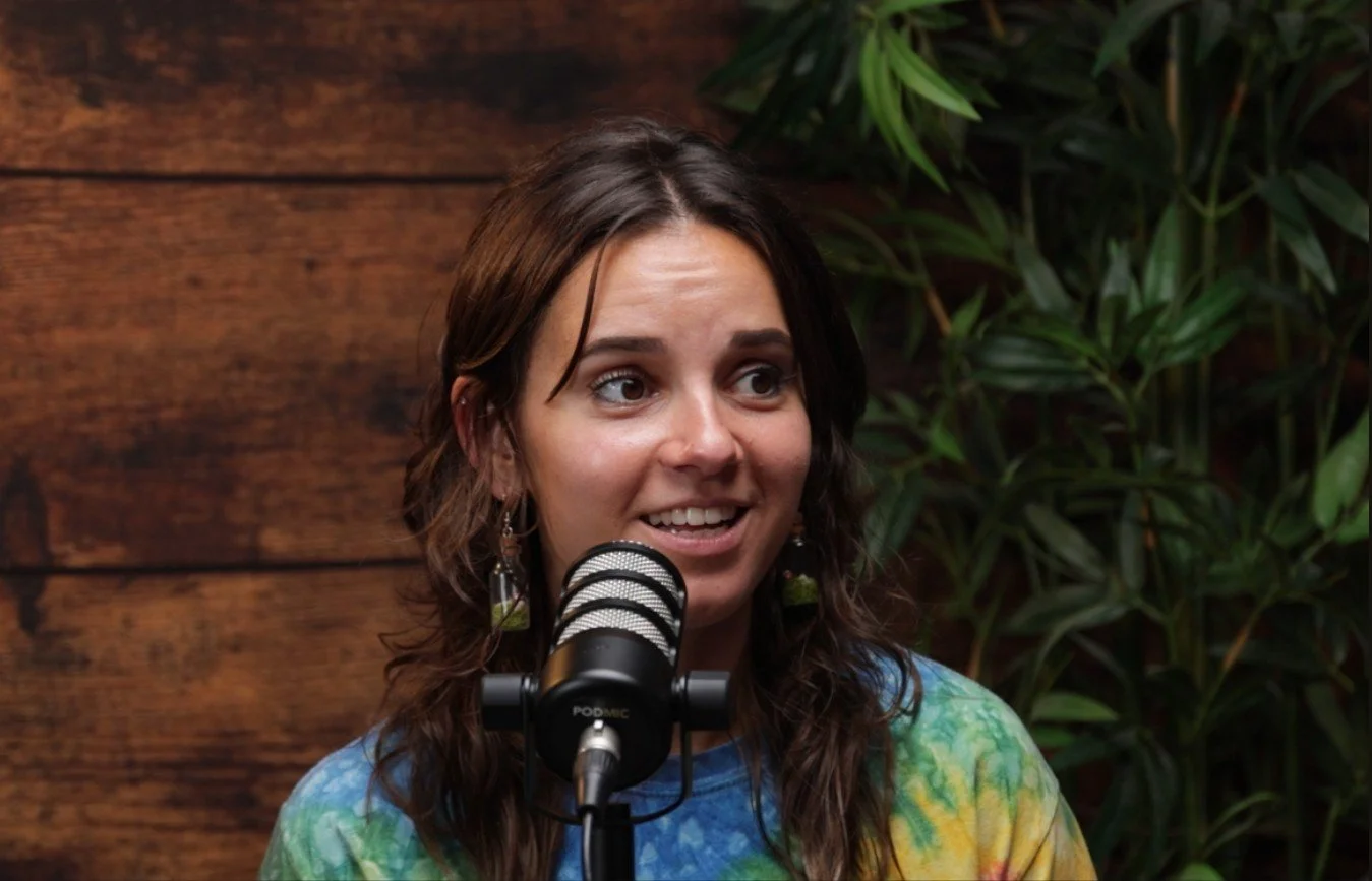 A woman with brown, curly hair is speaking into a microphone, wearing a colorful tie-dye shirt, with a wooden wall background and green plants behind her.